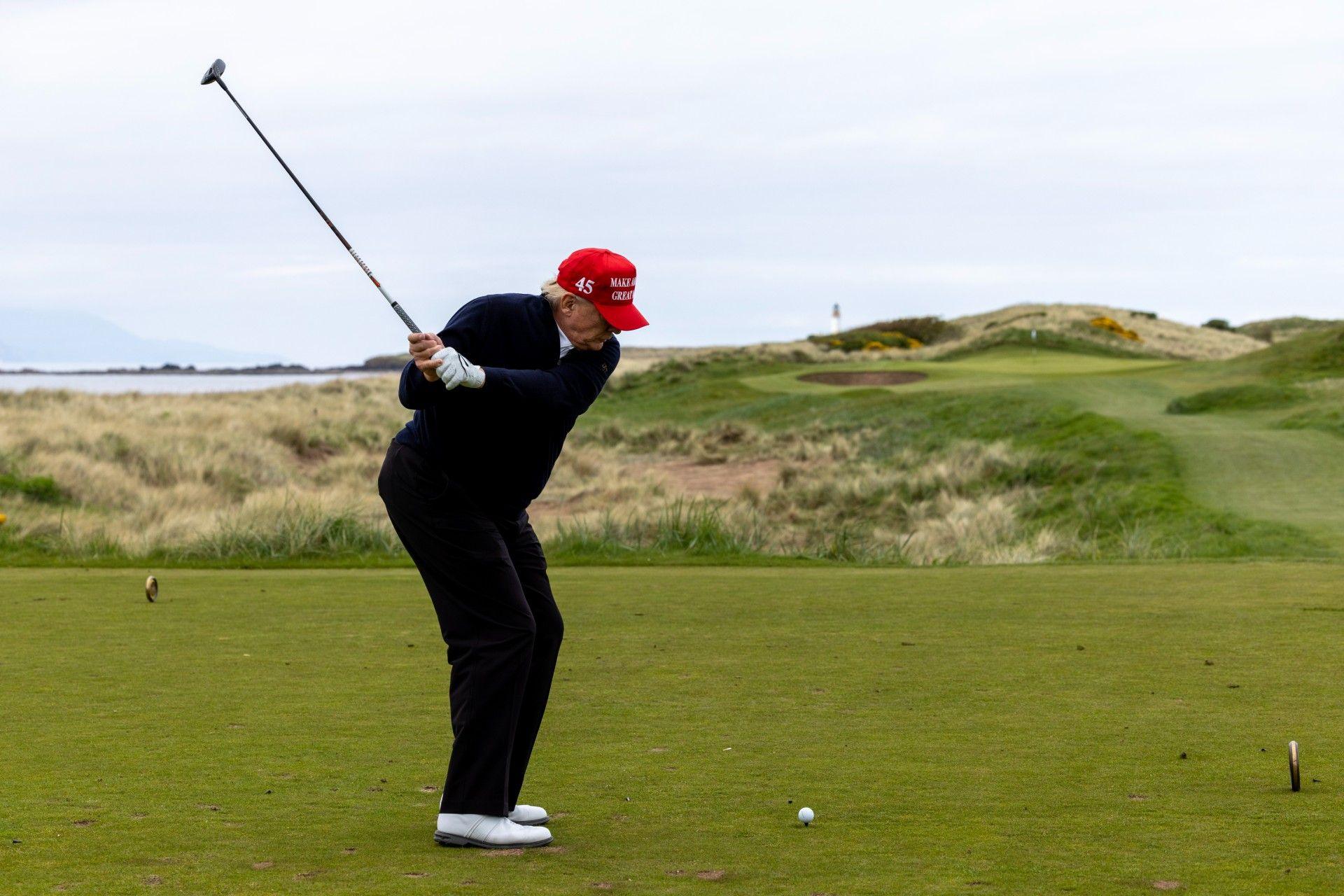 Trump takes a swing at his Turnberry golf course during a previous visit. He is wearing dark clothes, a red baseball cap and white golf shoes. The course is across sand dunes with the sea in the background