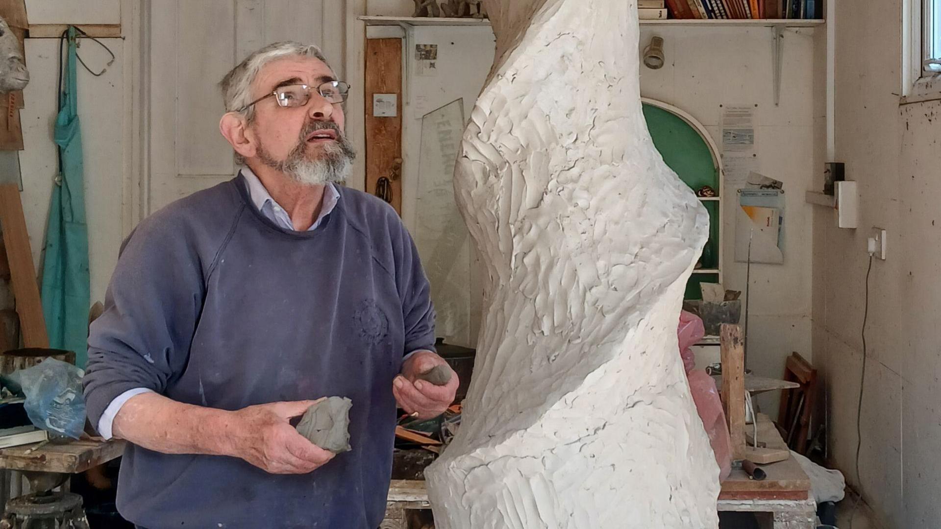 John working on the sculpture. He is looking up at a spiral made out of clay. He is wearing a blue jumper and glasses. He is in a studio.