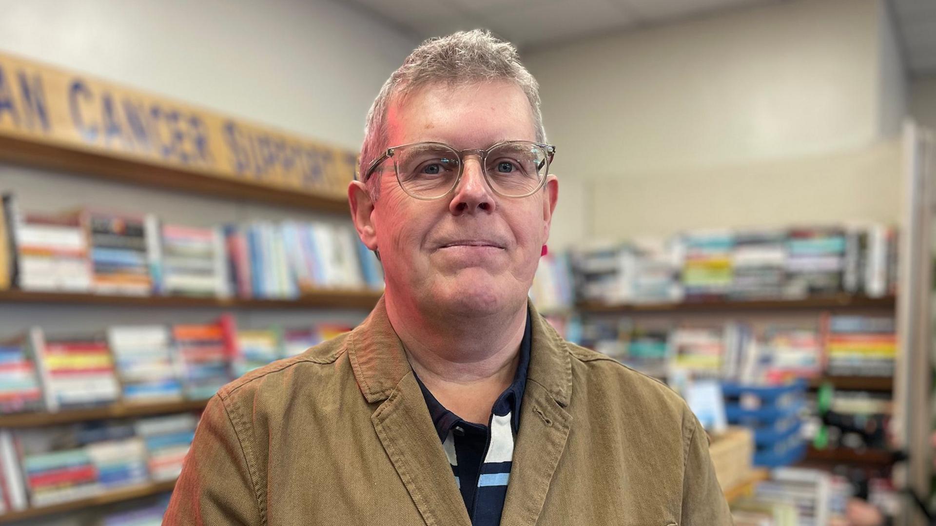 A grey haired bespectacled man in a brown jacket looks at the camera with second hand books in the background and a sign saying Arran Cancer Support