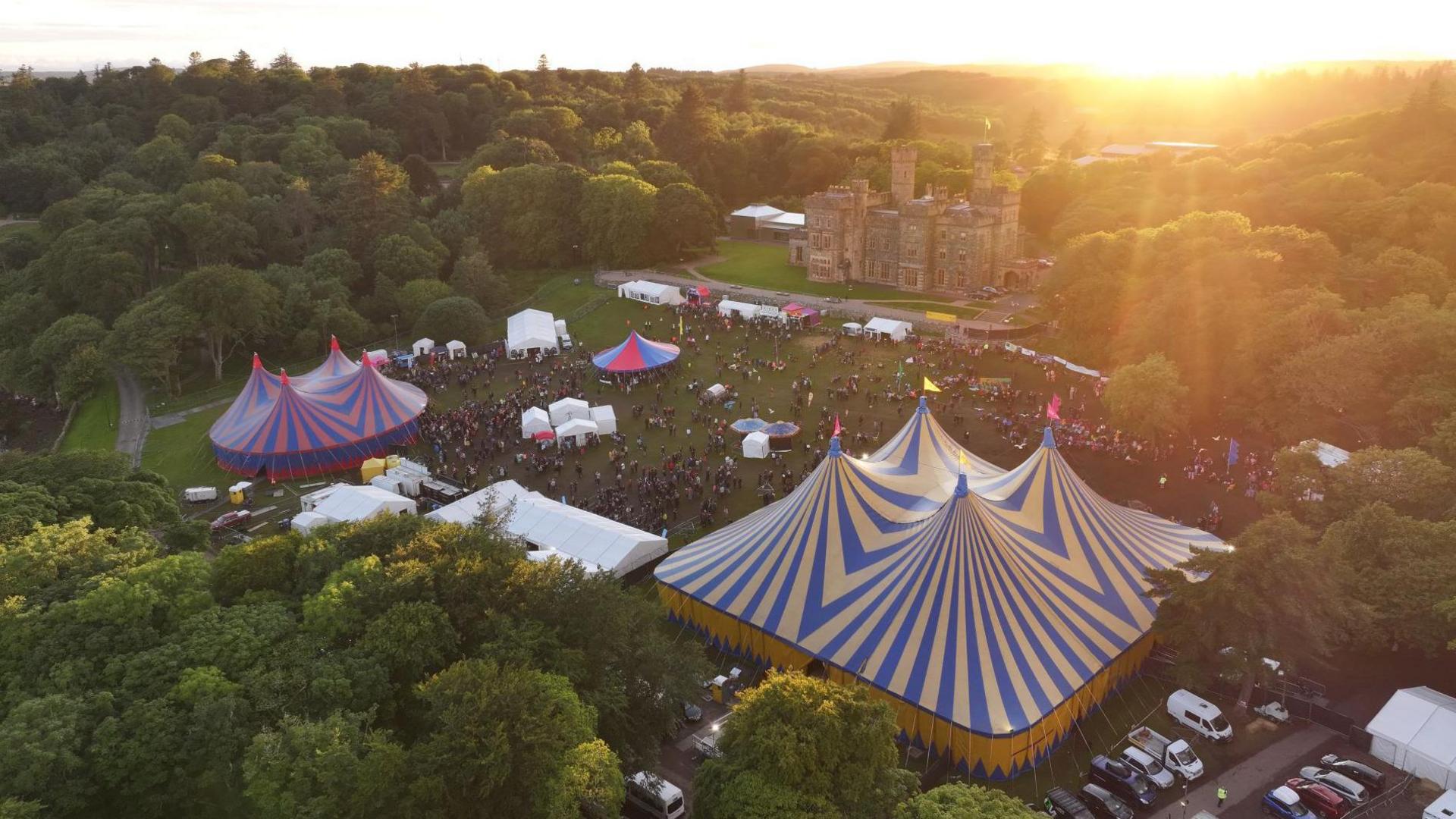 An aerial image of colourful circus big top-style tents on the festival site. There are people gathered in a grassy area between the tents. Lews Castle is in the background.