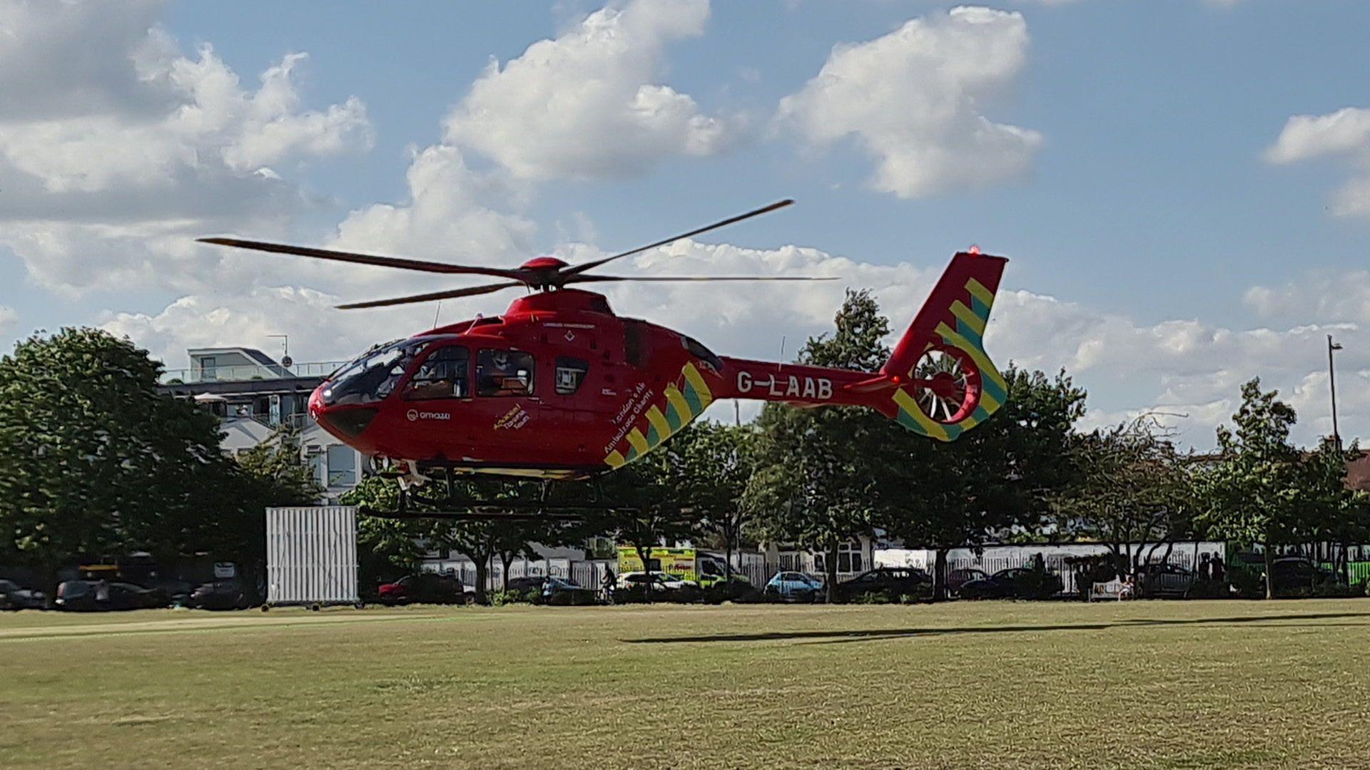 A red helicopter with yellow stripes on the tail hovers above the lawn of a large park, about 2 metres above it. You can see an ambulance on the road next to the park and cars parked alongside the fence. There are trees around the edge of the park.