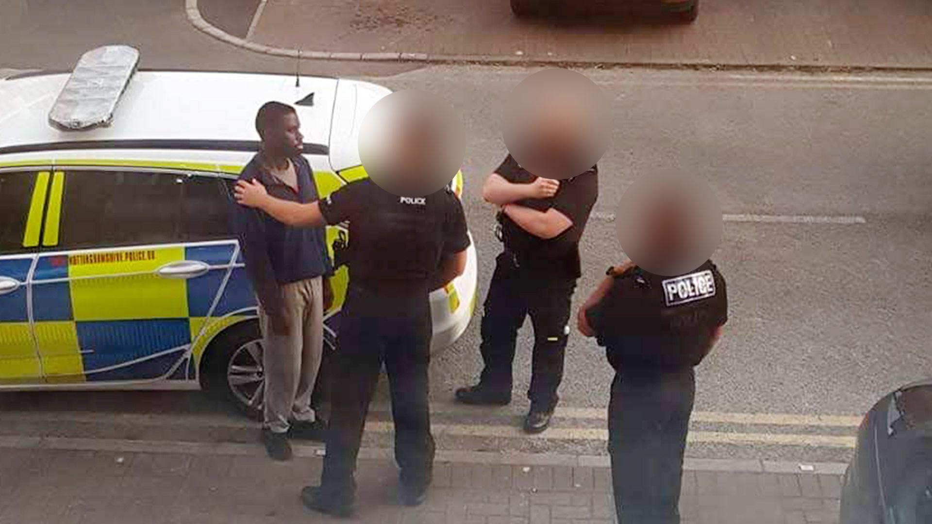 A photo taken from a window above of a police officer with his hand on Valdo Calocane's shoulder, next to a police car, with two more officers standing nearby