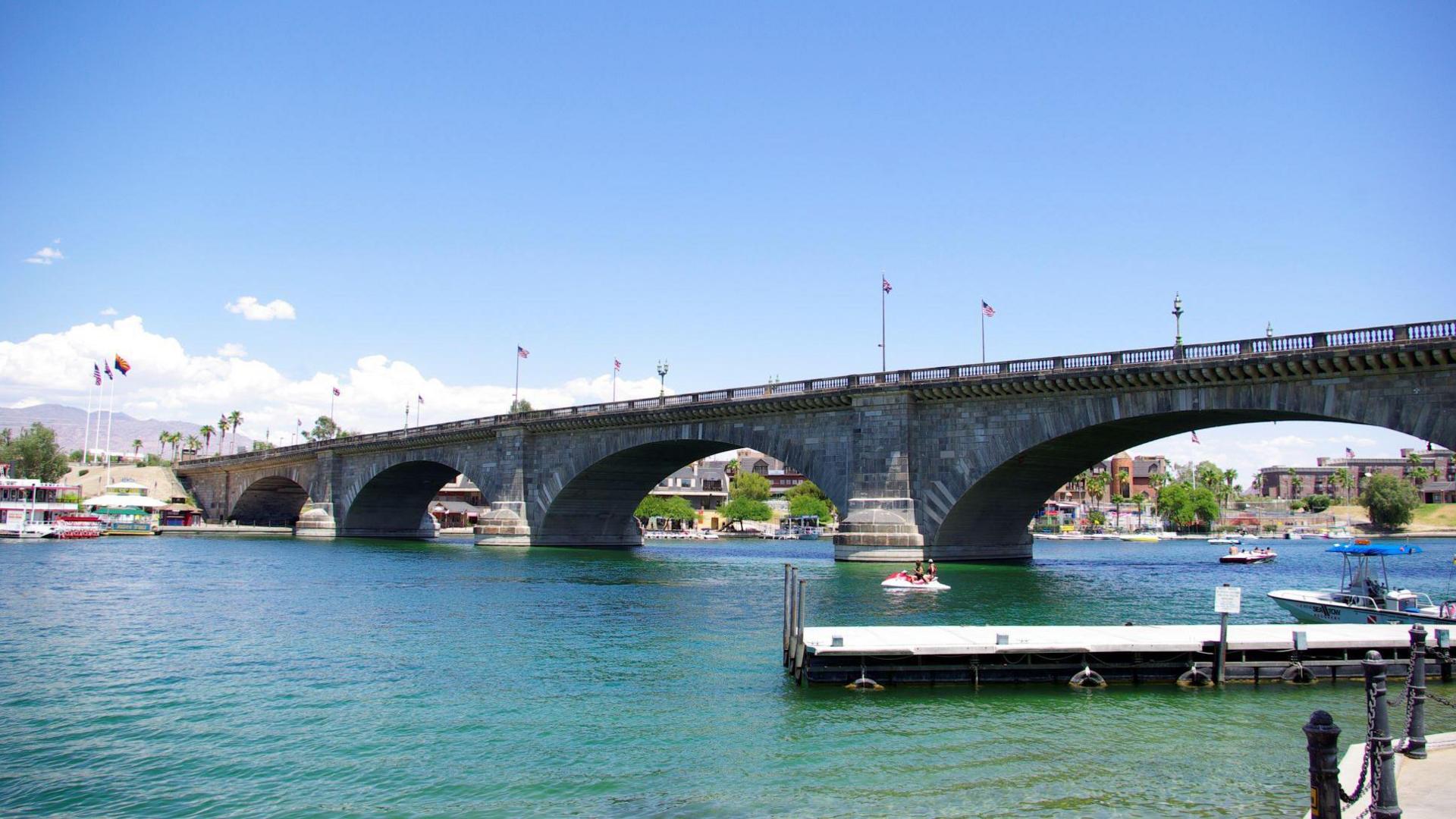 A modern-day image of the original bridge in Lake Havasu, Arizona - with US flags, a boat and a water scooter visible on the water 