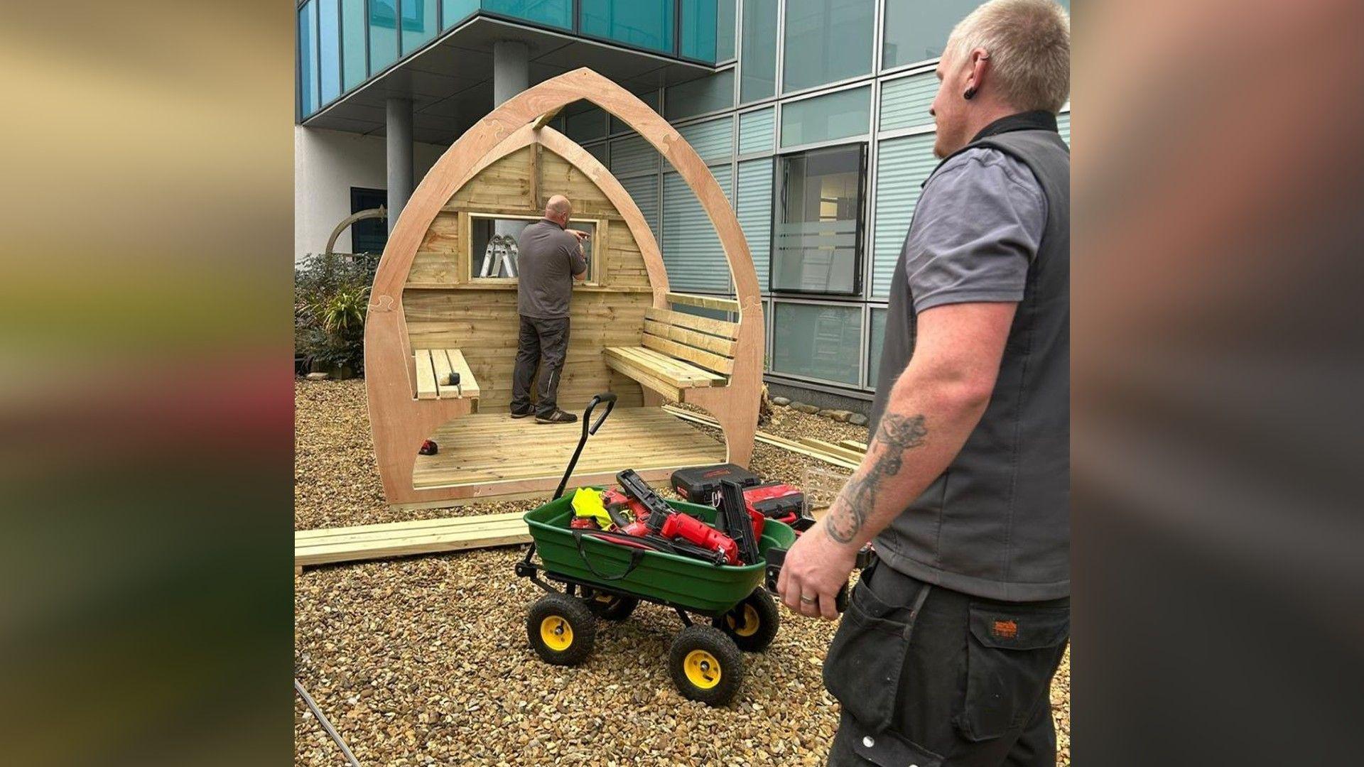 Workmen building a cone-shaped wooden pod. One of the men is standing in front of a green wheelbarrow