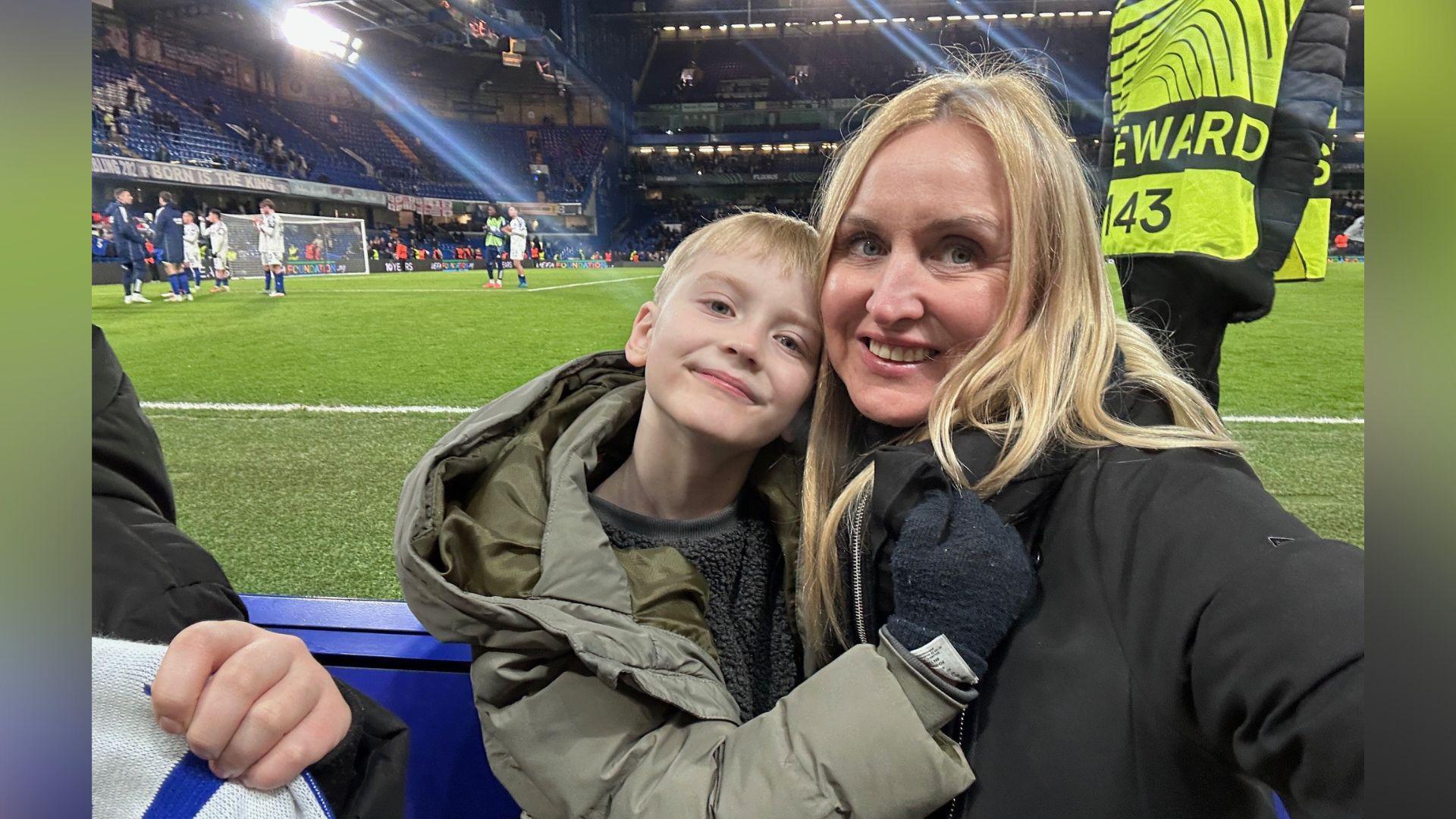 A boy with blond hair, wearing a khaki jacket, is hugging a woman with blonde hair and black jacket. They are watching a football match.