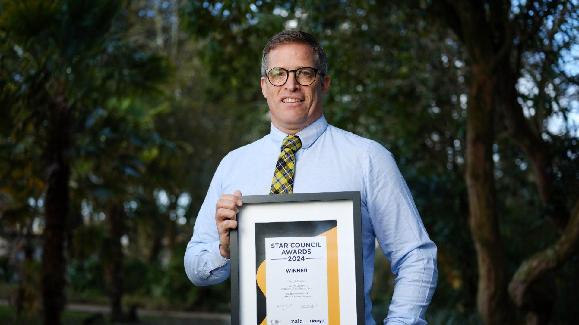 James Hardy, the town clerk for Penzance, holding a framed certificate. He has short dark hair, is wearing glasses, a yellow checked tie and a light full-sleeved shirt.