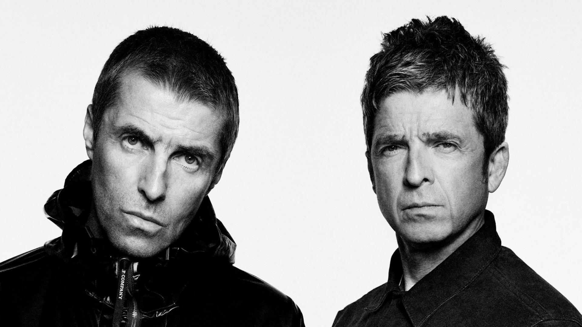 A 2024 black-and-white portrait of Liam and Noel Gallagher standing side by side against a plain background. Liam is wearing a shiny zip-up jacket and stares intently at the camera, while Noel wears a dark shirt and looks on with a serious expression. 