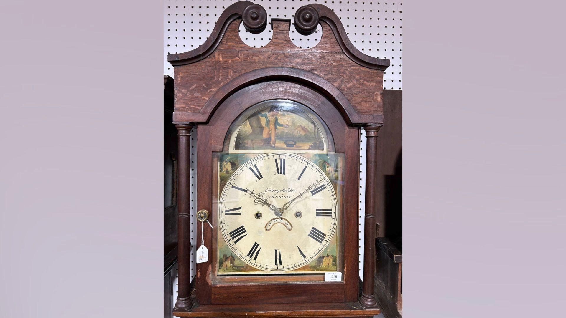 A brown wooden clock with a painted face that depicts houses and green grass and a boy above the face. The picture of the clock is on a light purple background.