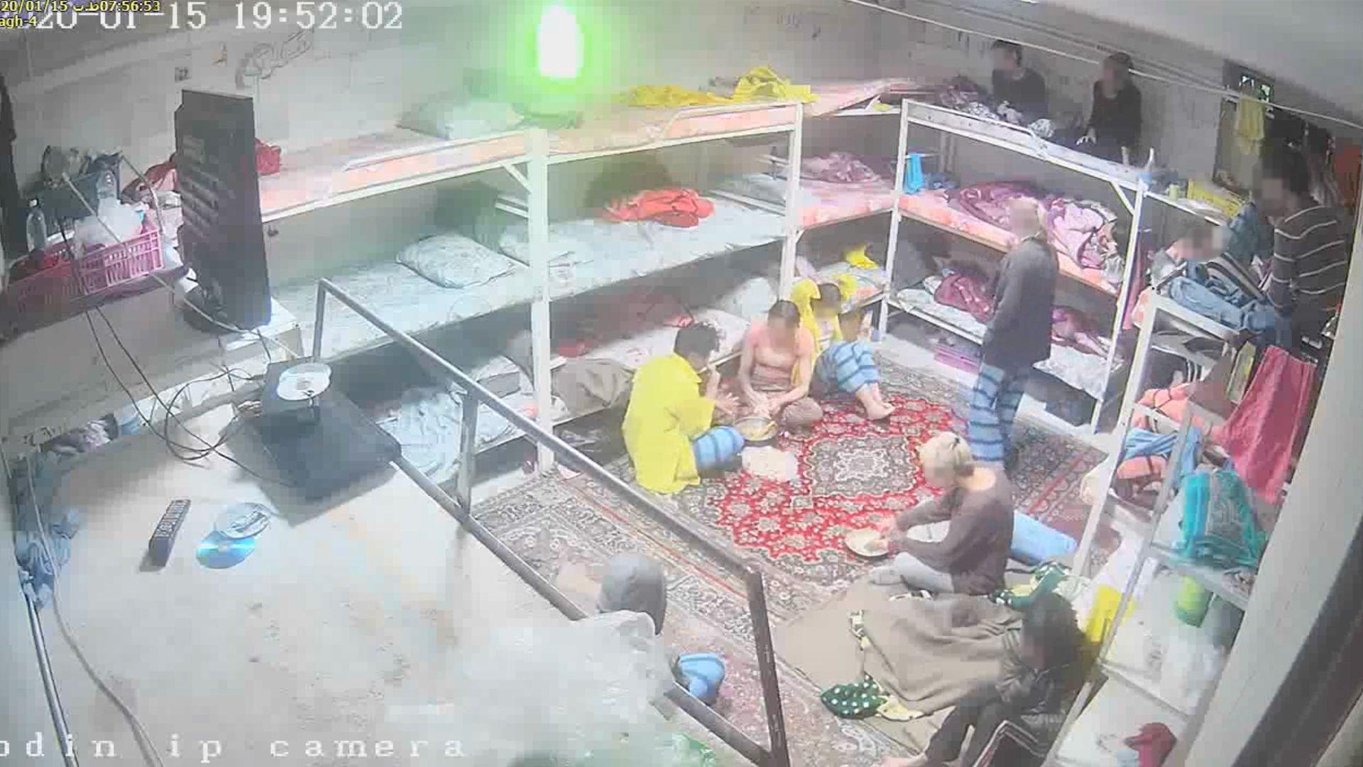 A frame from CCTV shows a room with bunk beds stacked three-high and end-to-end along the three walls that are visible. Approximately 18 beds are visible. Each bed has some bedding on it. People are sitting on some of the beds. Six people are sitting on the floor which is covered with a traditional patterned carpet. They are wearing casual clothing.