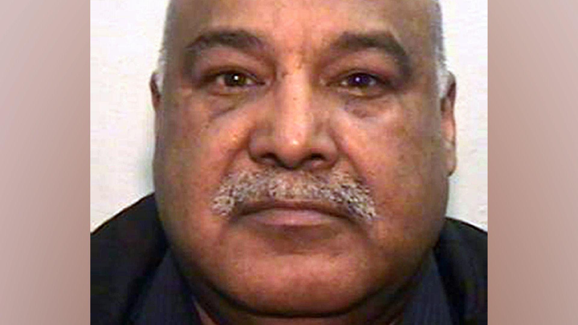 A mugshot of an Asian man in his 60s, with a grey moustache and bald head. He is looking into the camera.