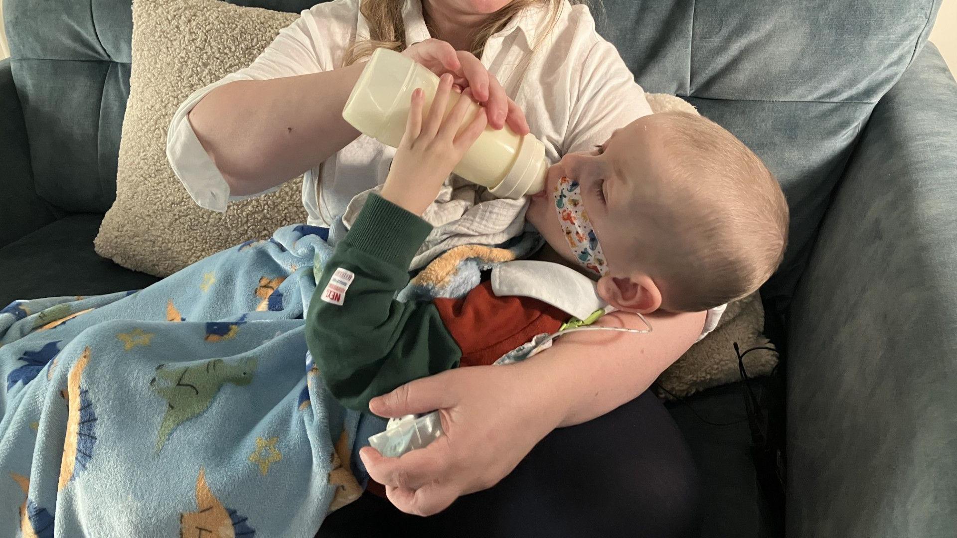 Deborah Rowe sitting on a grey sofa, with her son Jonah lying on her lap with a blue blanket. She is feeding him a bottle of milk. He has a nasogastric tube inserted and is lying against his mother's crooked arm.