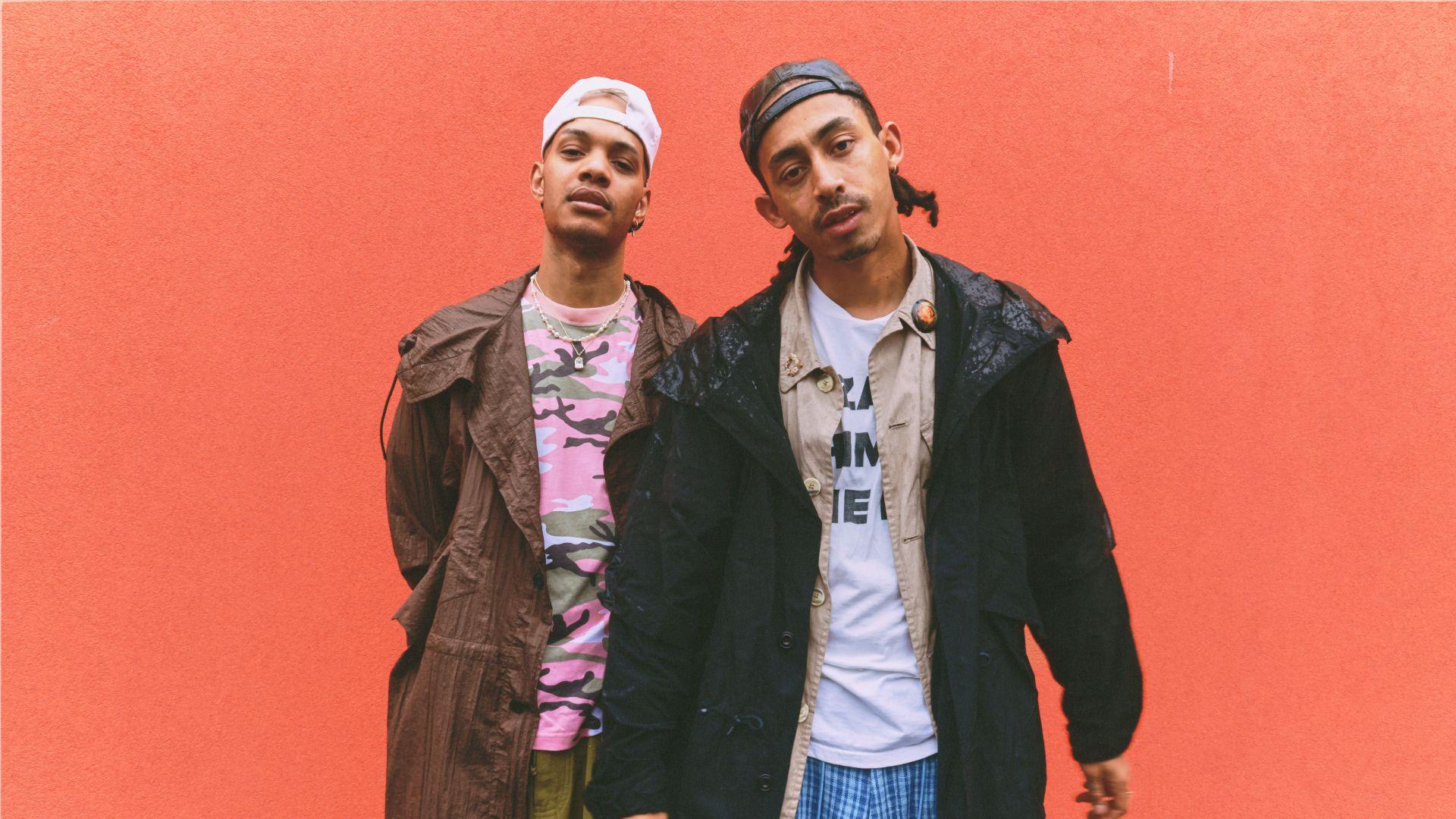 Harley Alexander-Sule and Jordan Stephens, aka Rizzle Kicks, look into the camera. Harley is wearing a pink camouflage t-shirt with a brown coat and backwards white baseball cap. Jordan is wearing a white t-shirt with a beige shirt and black coat over it, with a black baseball cap, also worn backwards. They are standing in front of a tomato-red background.