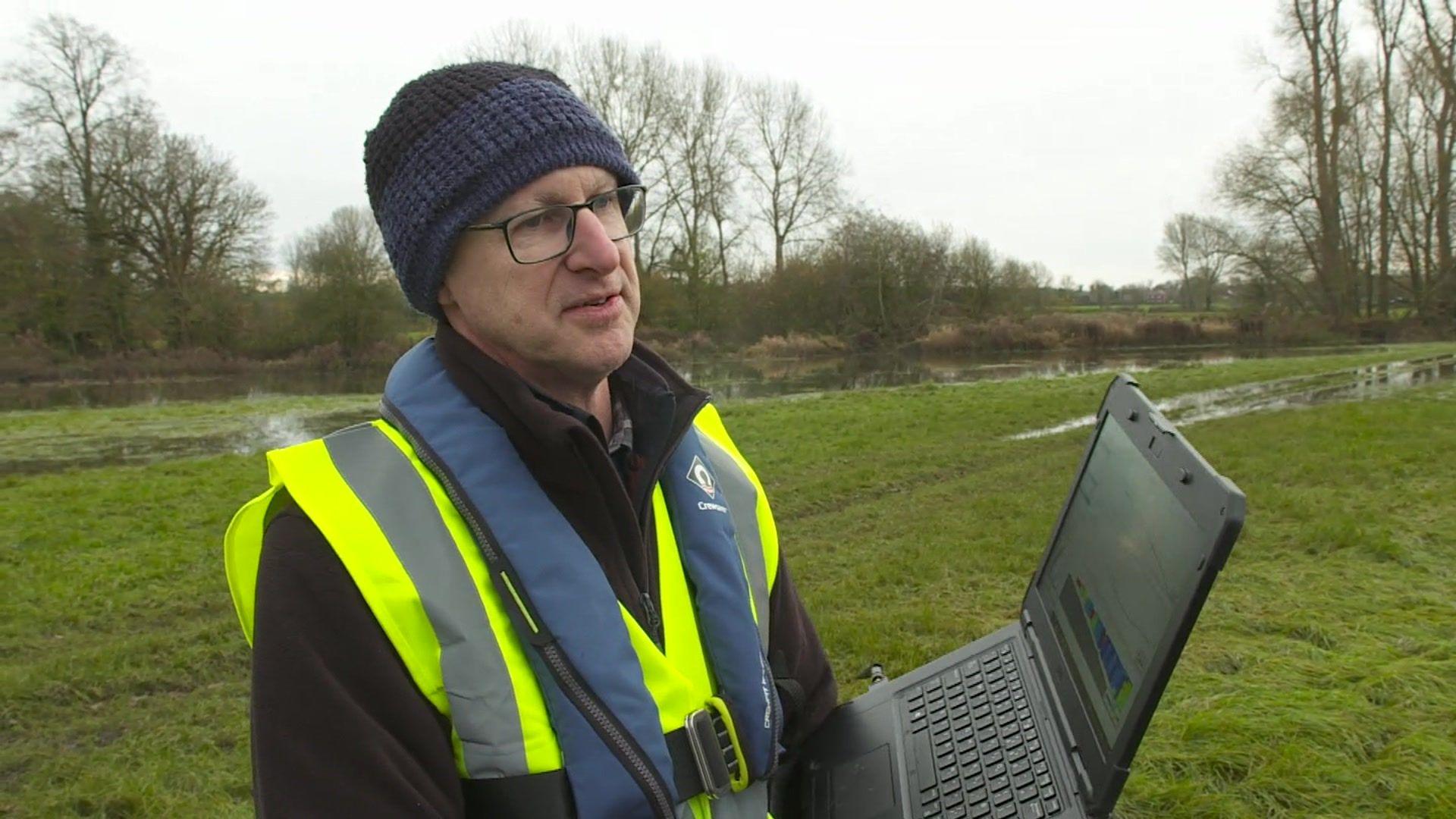 A picture of Nick Everard, who has a hi-vis jacket on and a black and blue woolly hat. He is holding a laptop computer in a waterlogged field.