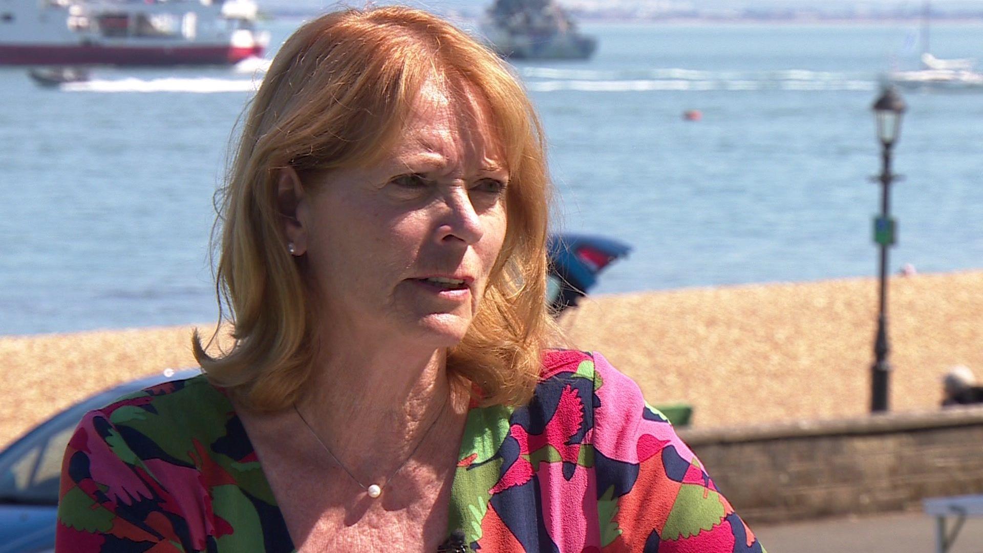 Isle of Wight resident Dee Oswald with red hair and wearing a multi-coloured top sitting in front of a beach in the background