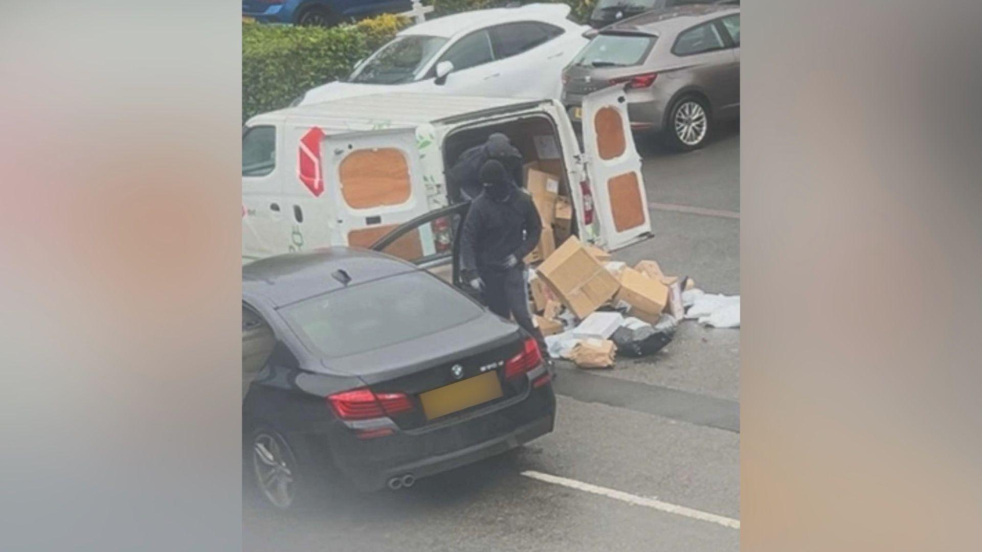 Two men wearing black clothing and black balaclavas, standing between a DPD delivery van which has its rear doors open and parcels spilling from it, and a black BMW.