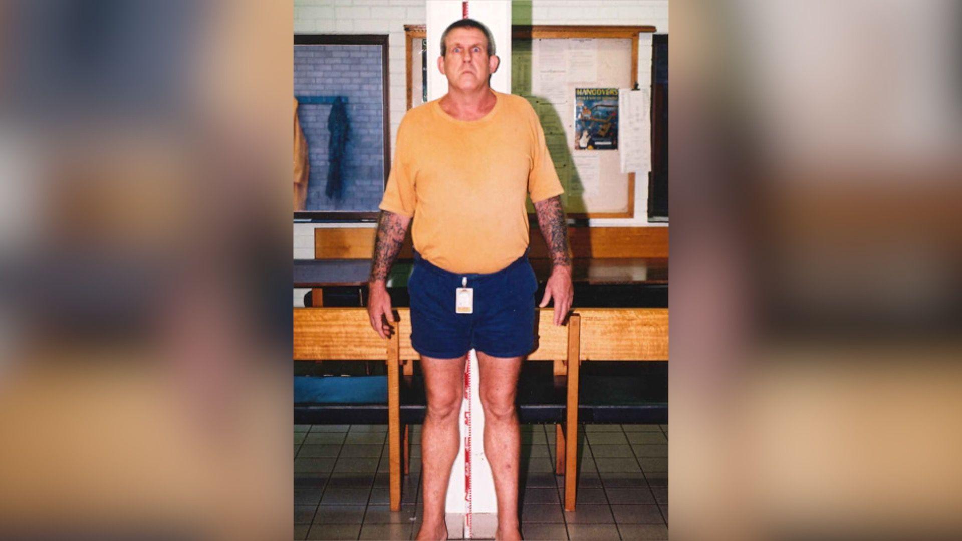 A tall man in a yellow t-shirt and blue short stands against a measuring tape in an office