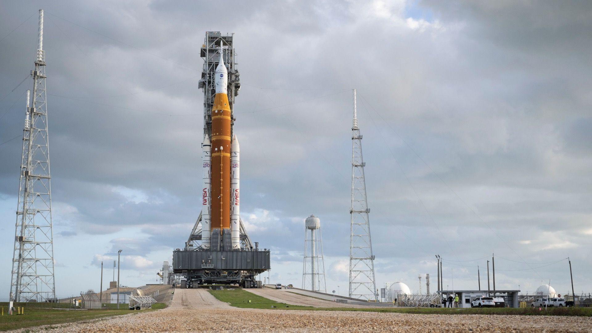 A large orange rocket, secured to a mobile launcher, being transported up small slope by a big tank-like vehicle on a cloudy day.