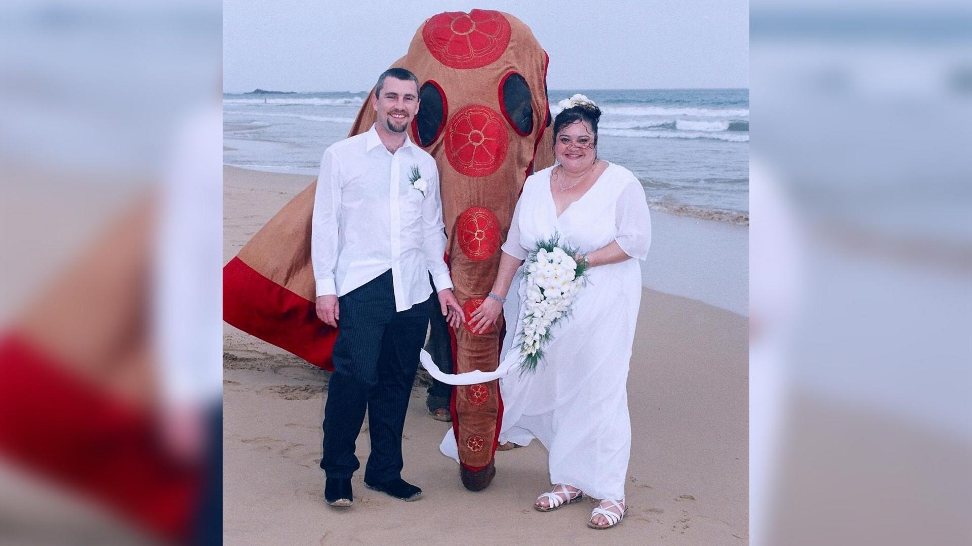 A man in a white shirt and dark trousers, and a woman wearing a white wedding dress and holding a bunch of flowers, stand on a beach in front of an elephant wearing a decorated sheet.