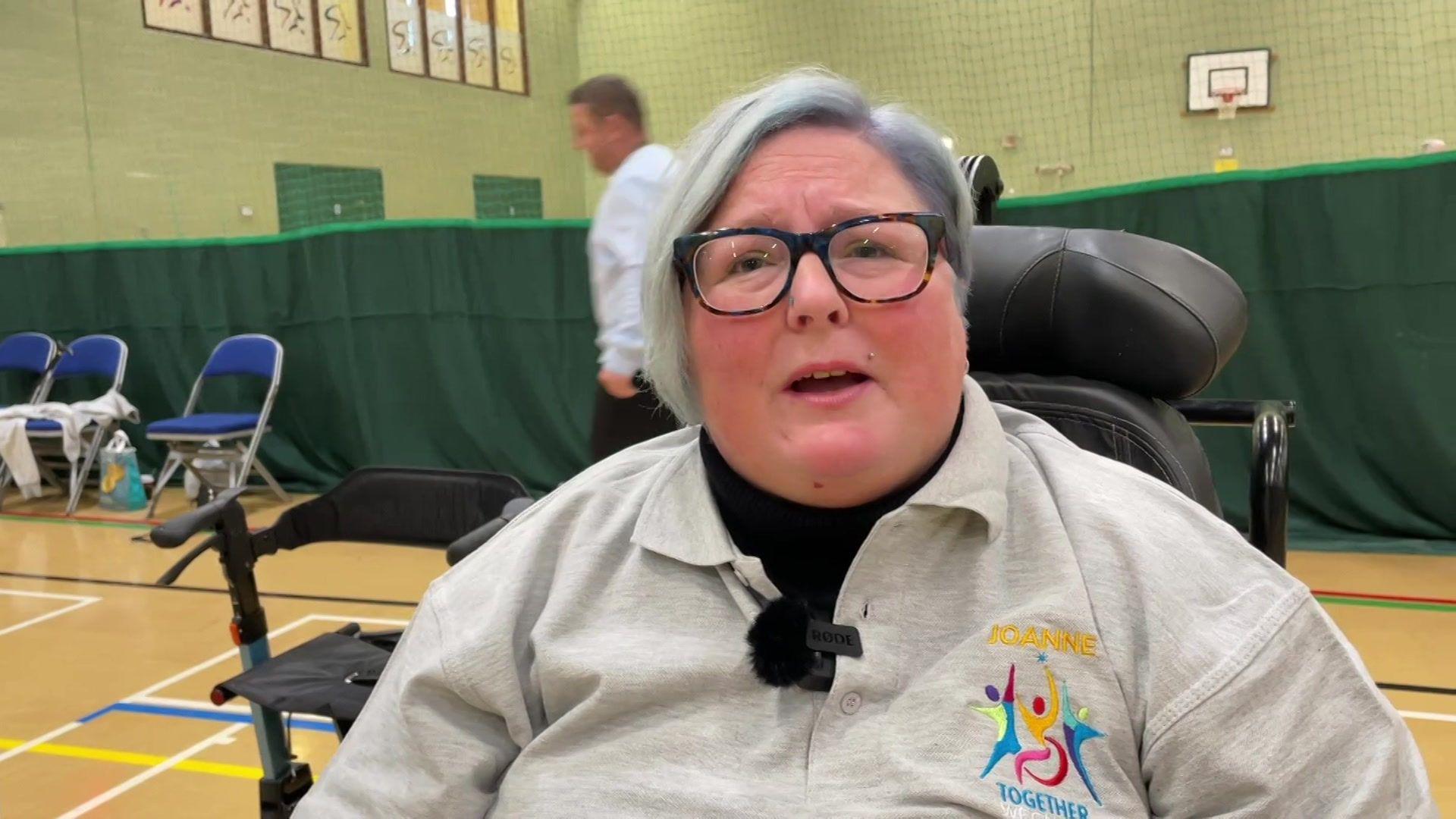 Wheelchair user Joanne Quinnell inside a sports hall. She has short grey hair dark rimmed glasses and is wearing a grey sports top.