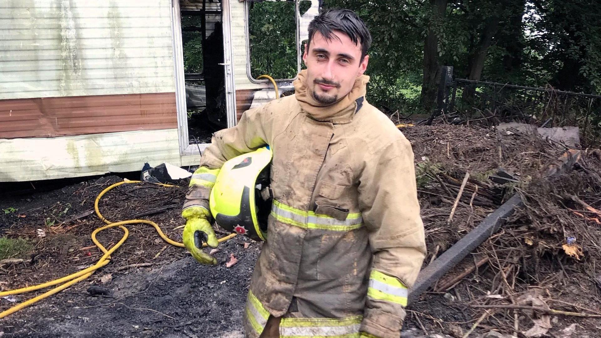 Oliver Dent wearing a firefighter's uniform with a yellow helmet tucked under one arm and standing in front of a burnt caravan.