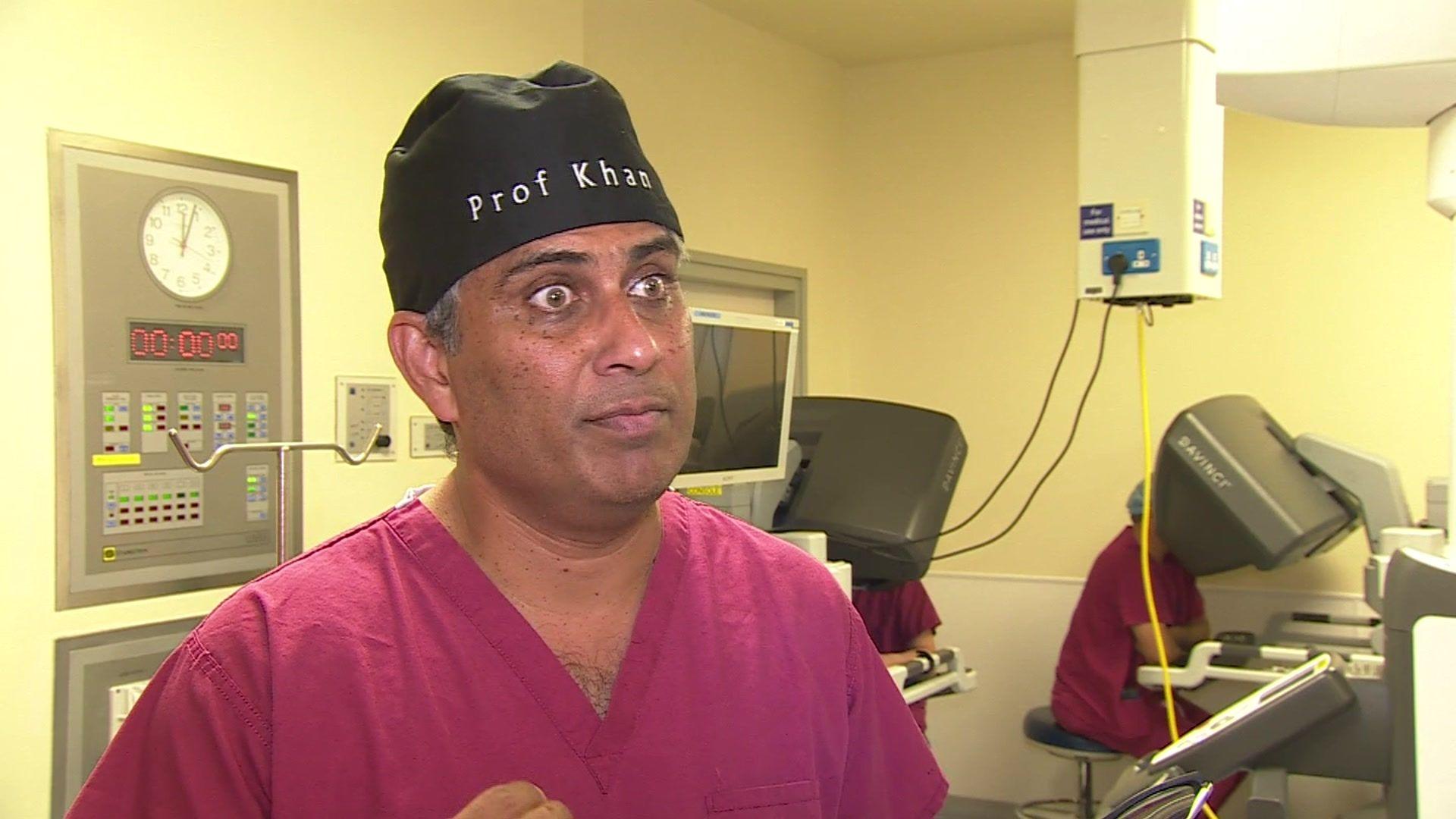 Prof Jim Khan in maroon medical dress, standing in an operating theatre. He is wearing a black head covering with the name 'Prof Khan' written in white