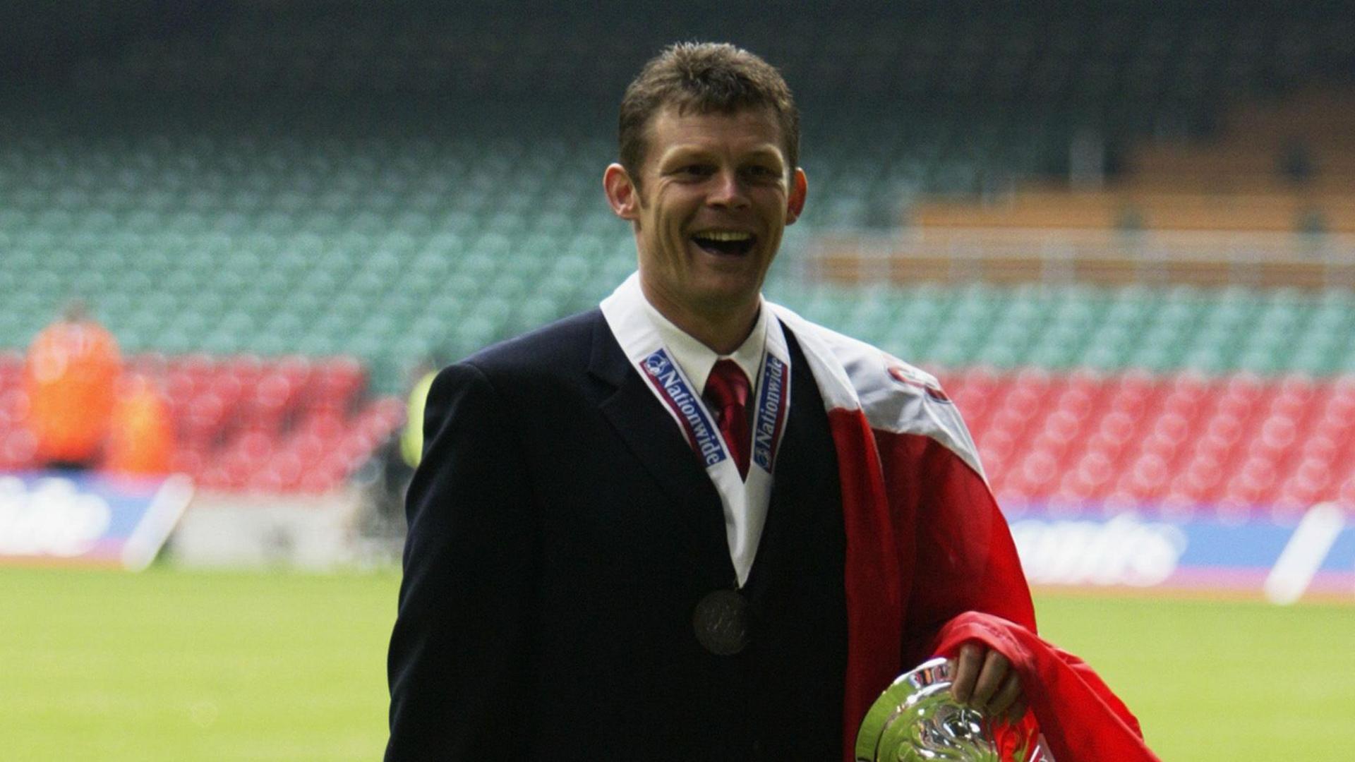 Steve Cotterill walking on the pitch at Wembley in 2002 holding the Division Three trophy with a scarf draped over his shoulder