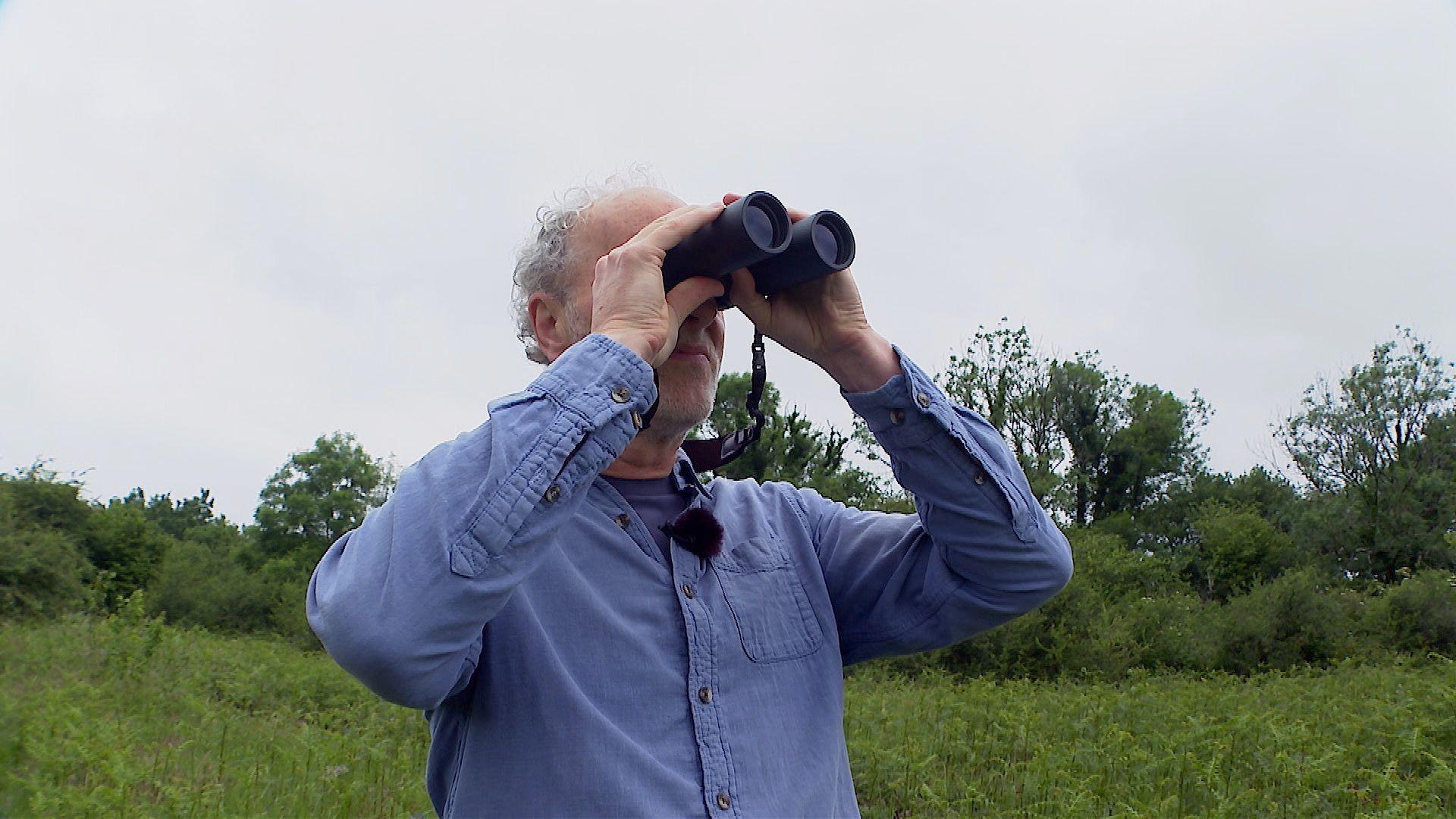 Charlie Elder is pictured though his face is not really visible as he is holding black binoculars to his eyes. He is wearing a blue shirt over a blue T-shirt and he has curly grey hair.