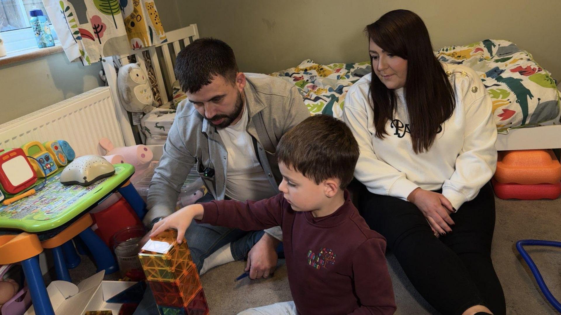 Father and mother sit on the floor of their son's bedroom and play with him, building bricks. A bed is seen in the background with a colourful dinosaur bedcover on it, and there is a green, orange and blue plastic table and chair set in the room. 