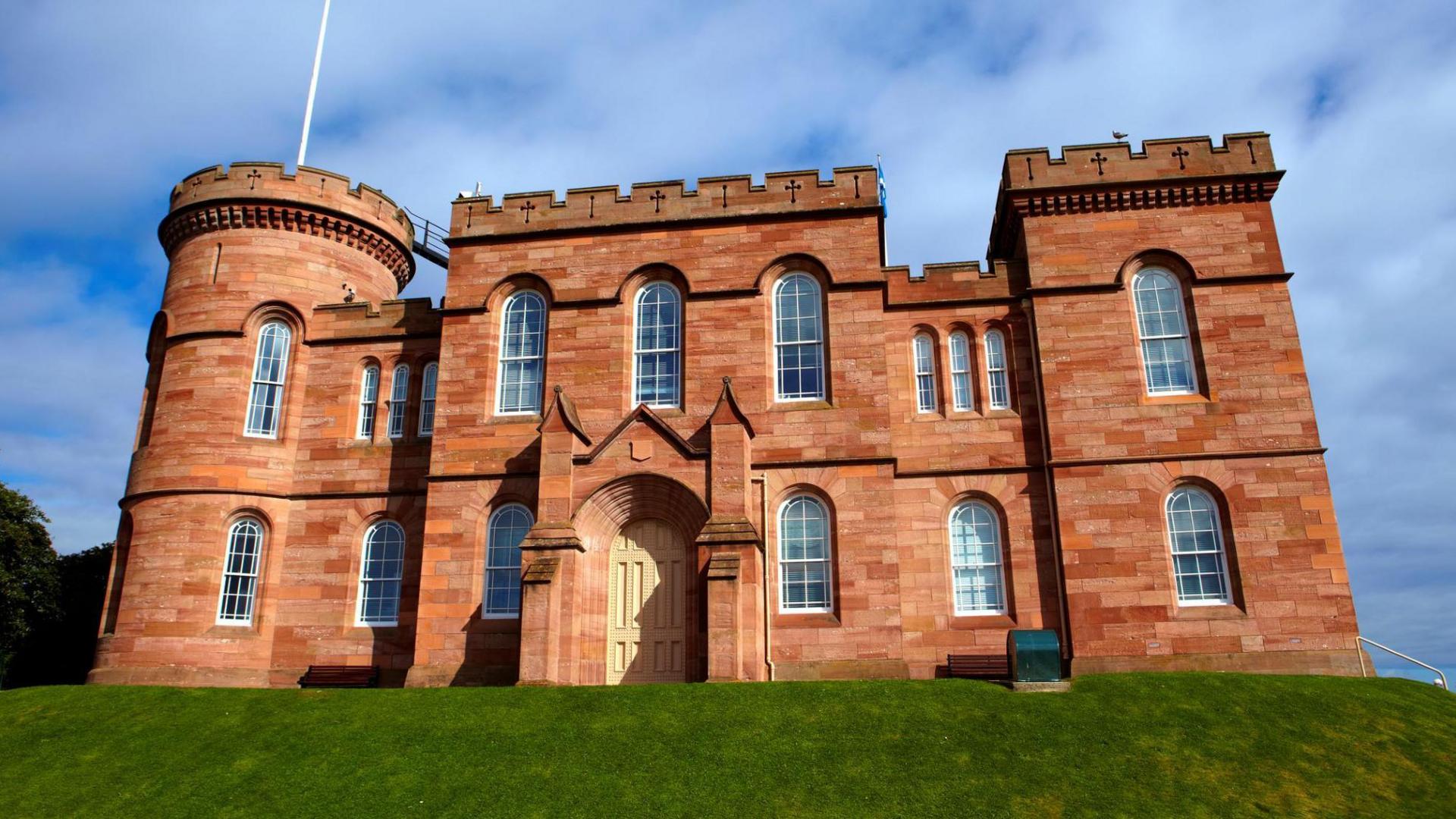 A view looking up at Inverness Castle. It is a sandstone building with turrets and battlements. There is an area of cut grass in front of the castle.