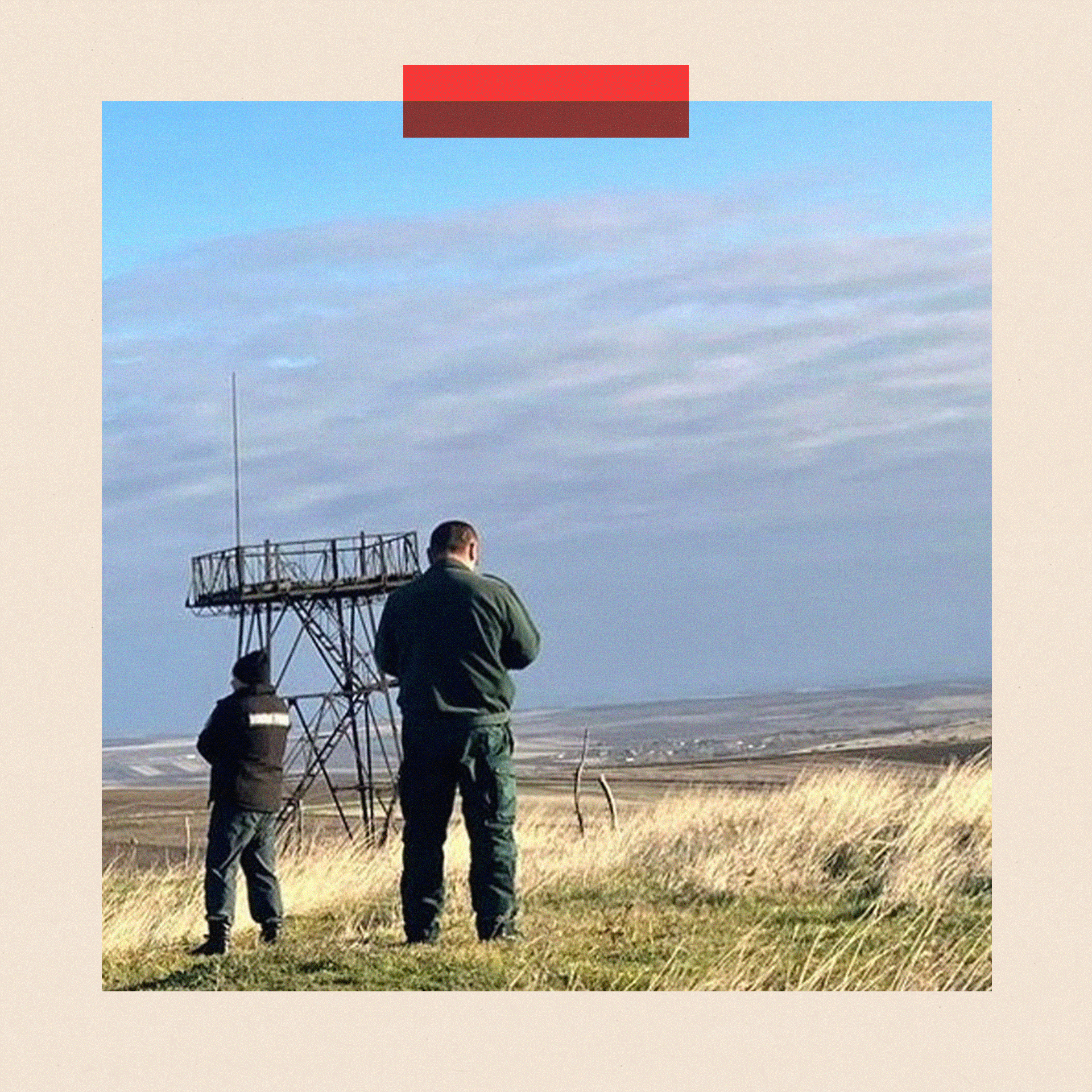 Two men stand with their backs to camera in a field with what appears to be a lookout tower behind them.