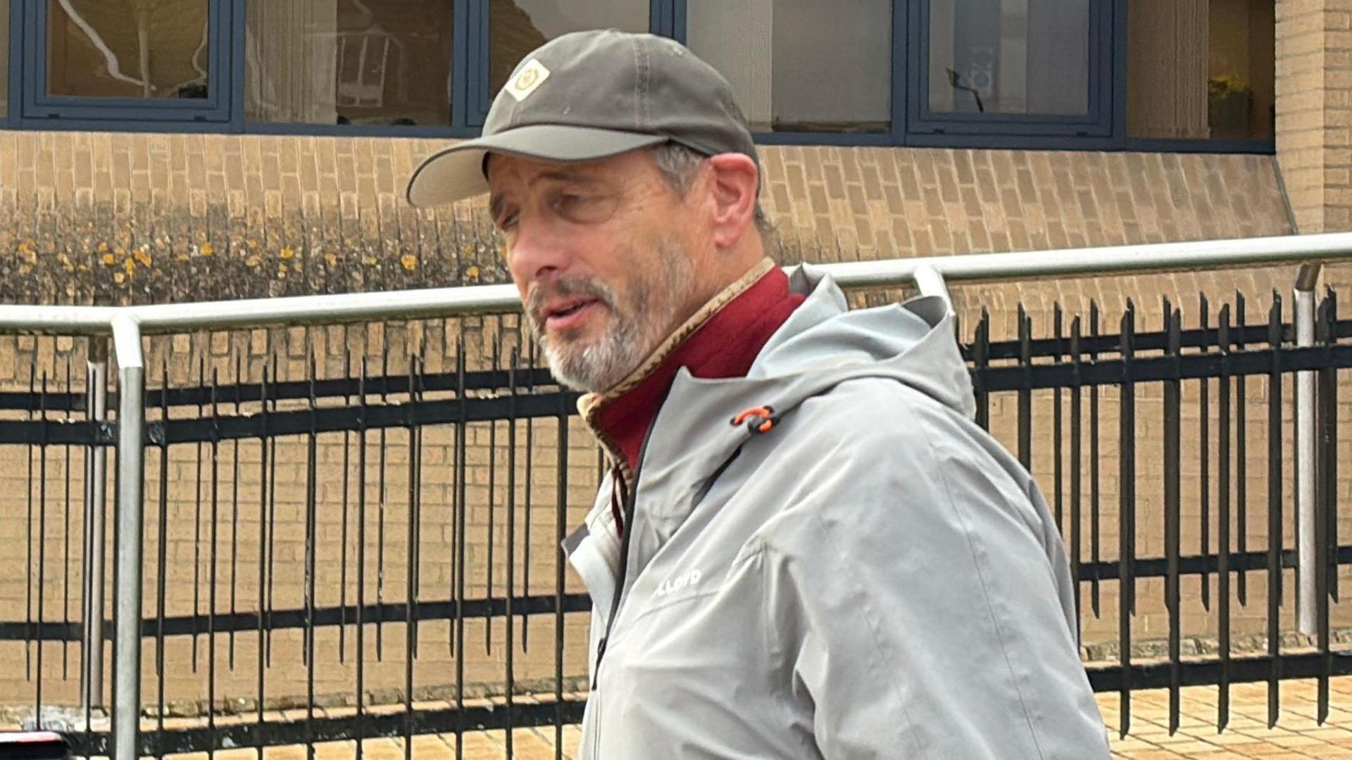 A man walking in front of a yellow brick building and railings. He wears a dark grey baseball cap and  light grey rain jacket. A burgundy top can be seen underneath. He has a grey beard and mustache which is kept short.