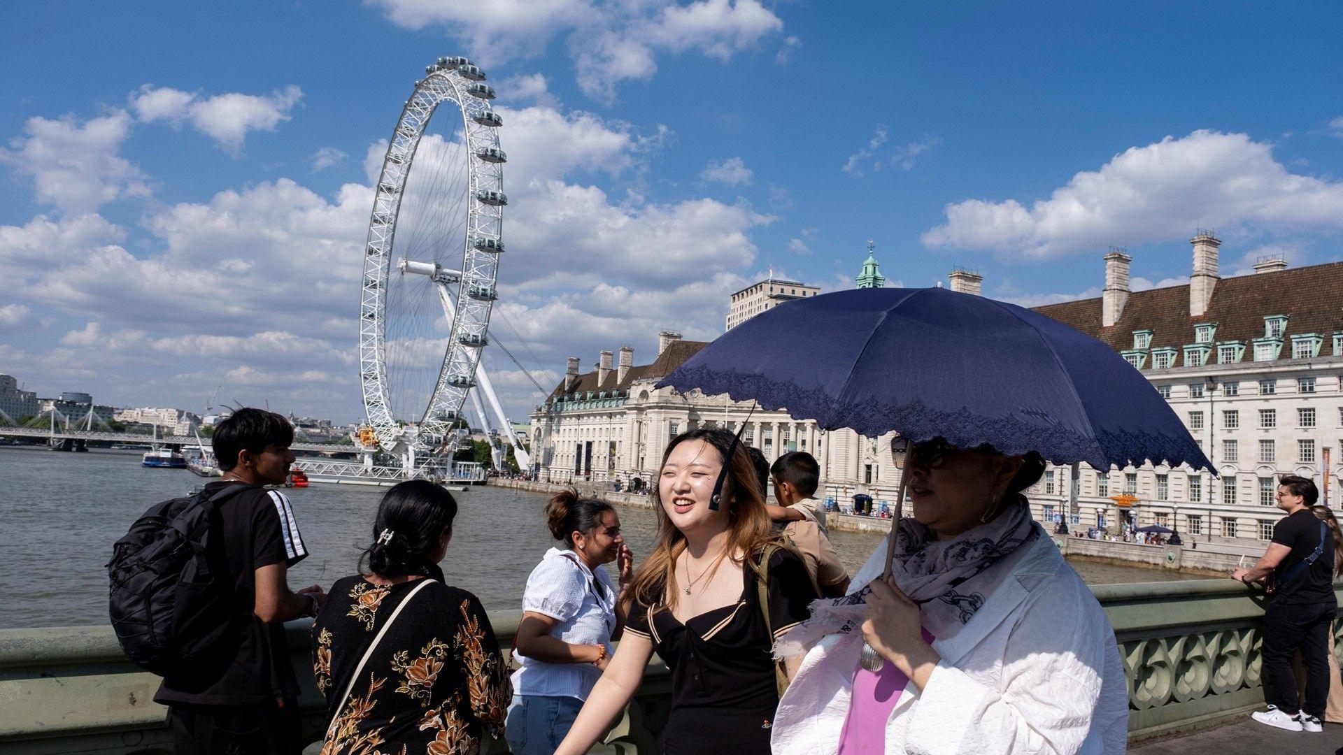 People walk on a bridge over the Thames in London using an umbrella for shade with strong sunshine overhead.