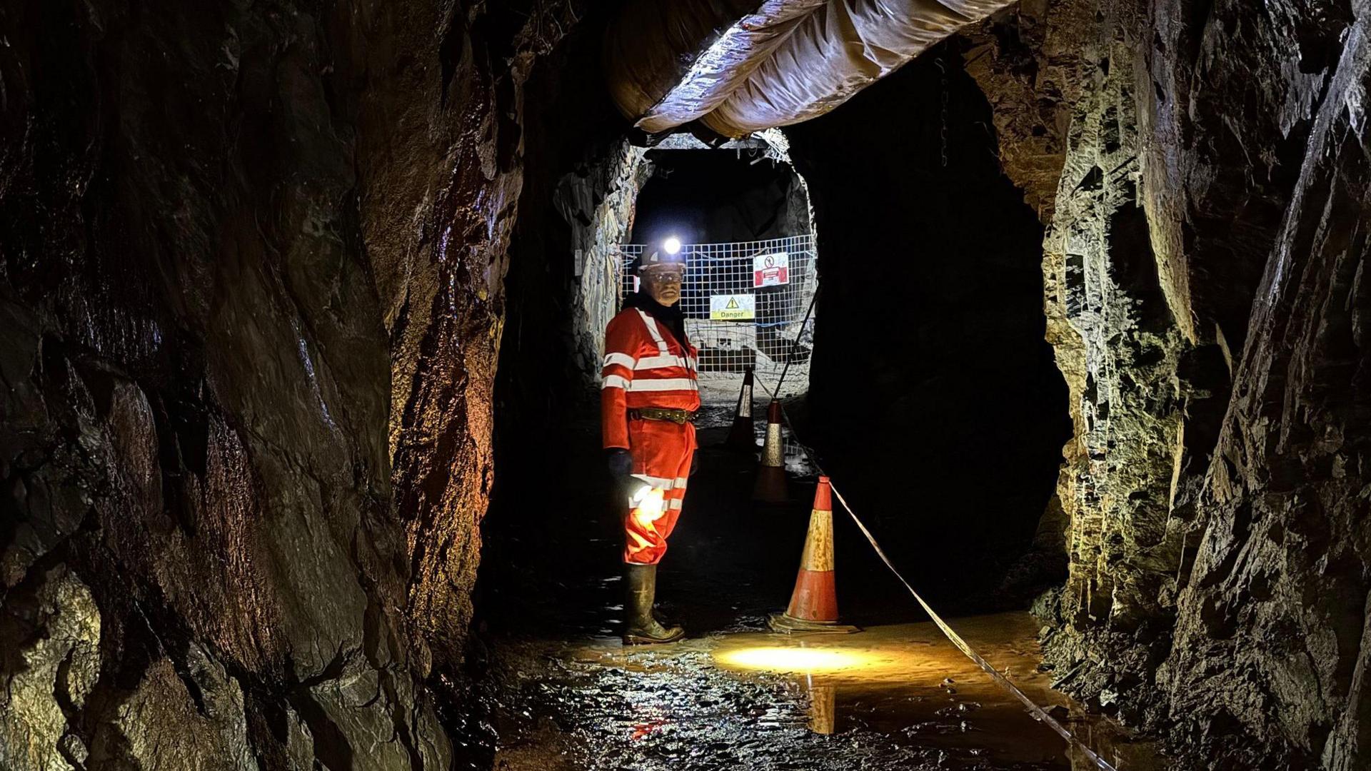 A mine worker in orange high viz clothing, a hard hat with a lamp on it and boots stands in a mine tunnel with dark grey walls and water on the ground 