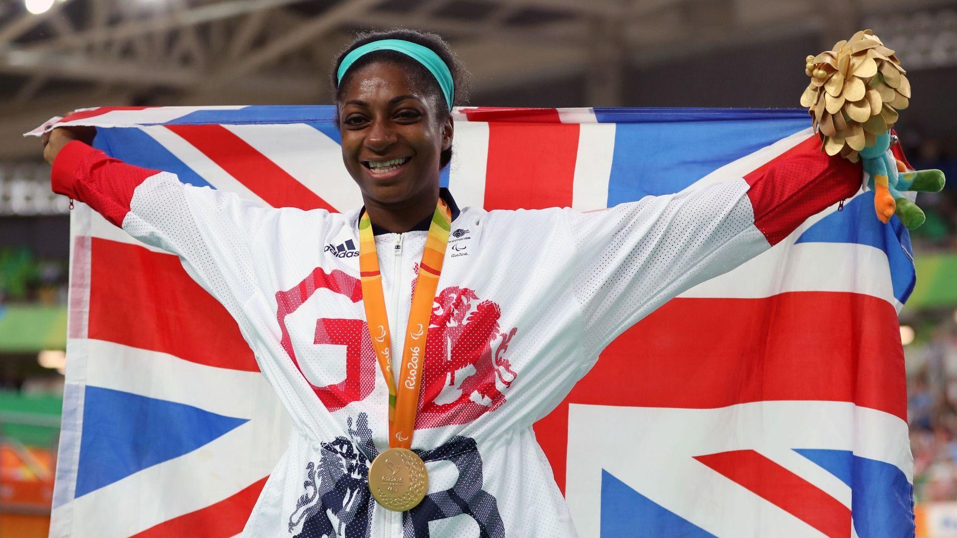 Kadeena Cox wearing a Team GB tracksuit as she holds a Union Jack in a sports stadium. She has a gold medal around her neck and is holding a bunch of flowers.