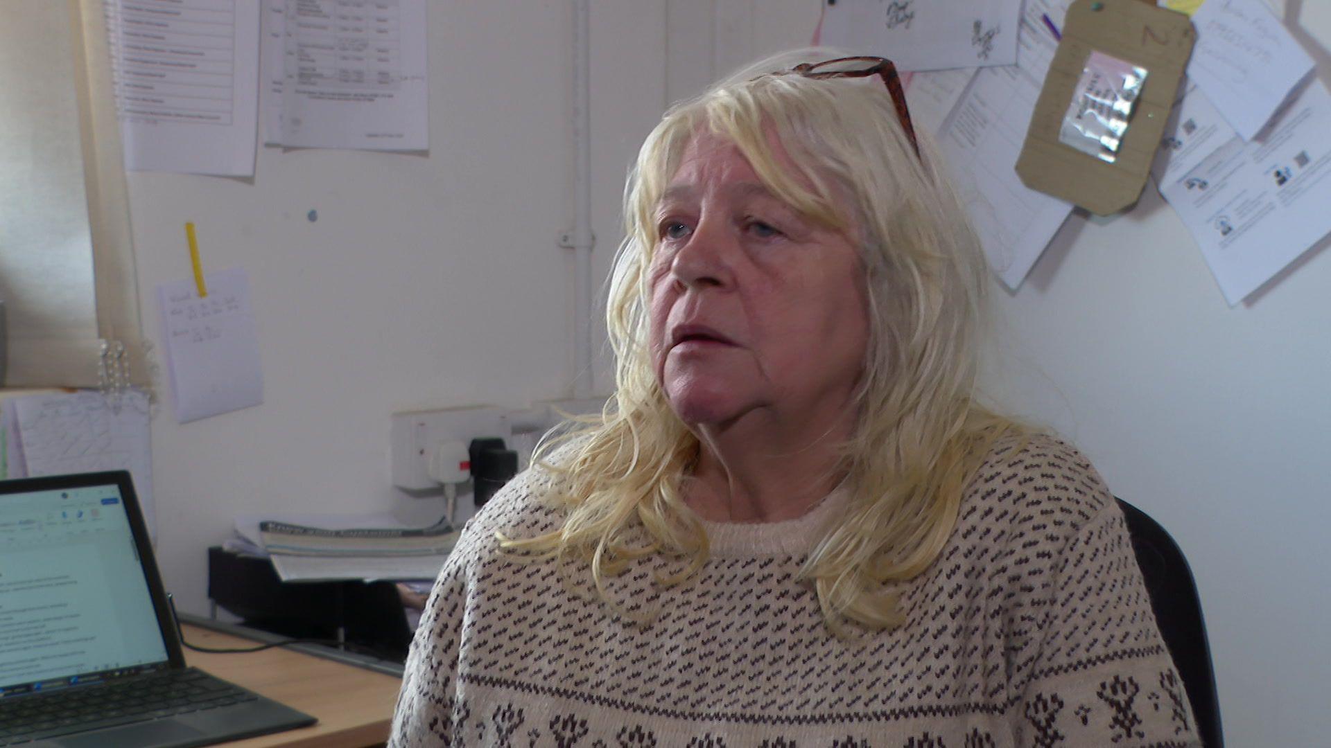 Angela Buss, wearing a white patterned jumper, sits in an office and speaks to the camera