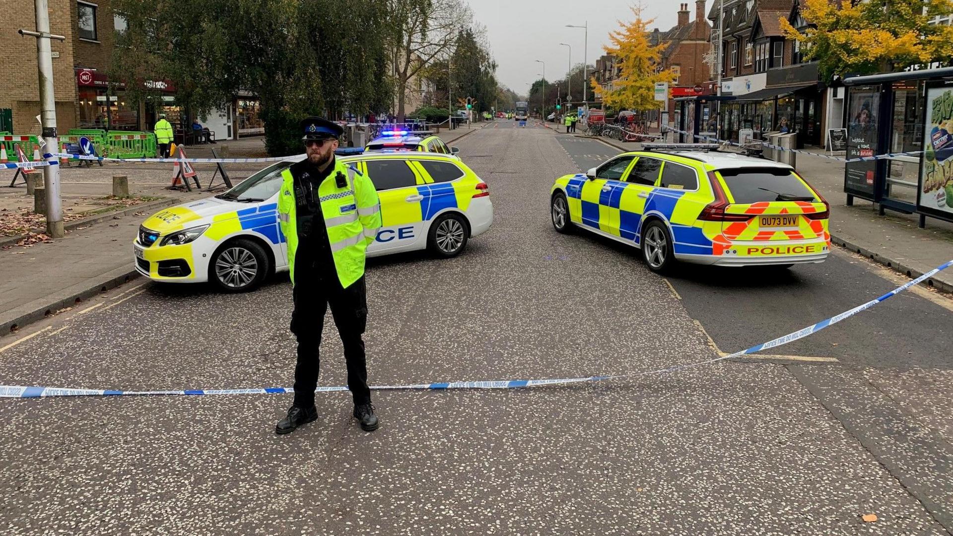 A police officer stands in the centre of Banbury Road. A cordon behind him stretches across the road and off into the distance on both sides. Three police cars are also in the road.