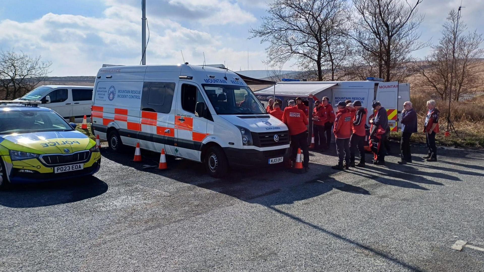 A police vehicle parked next to mountain rescue vehicles in a car park. A huddle of people in orange jackets can be seen gathered behind one of the vans. 