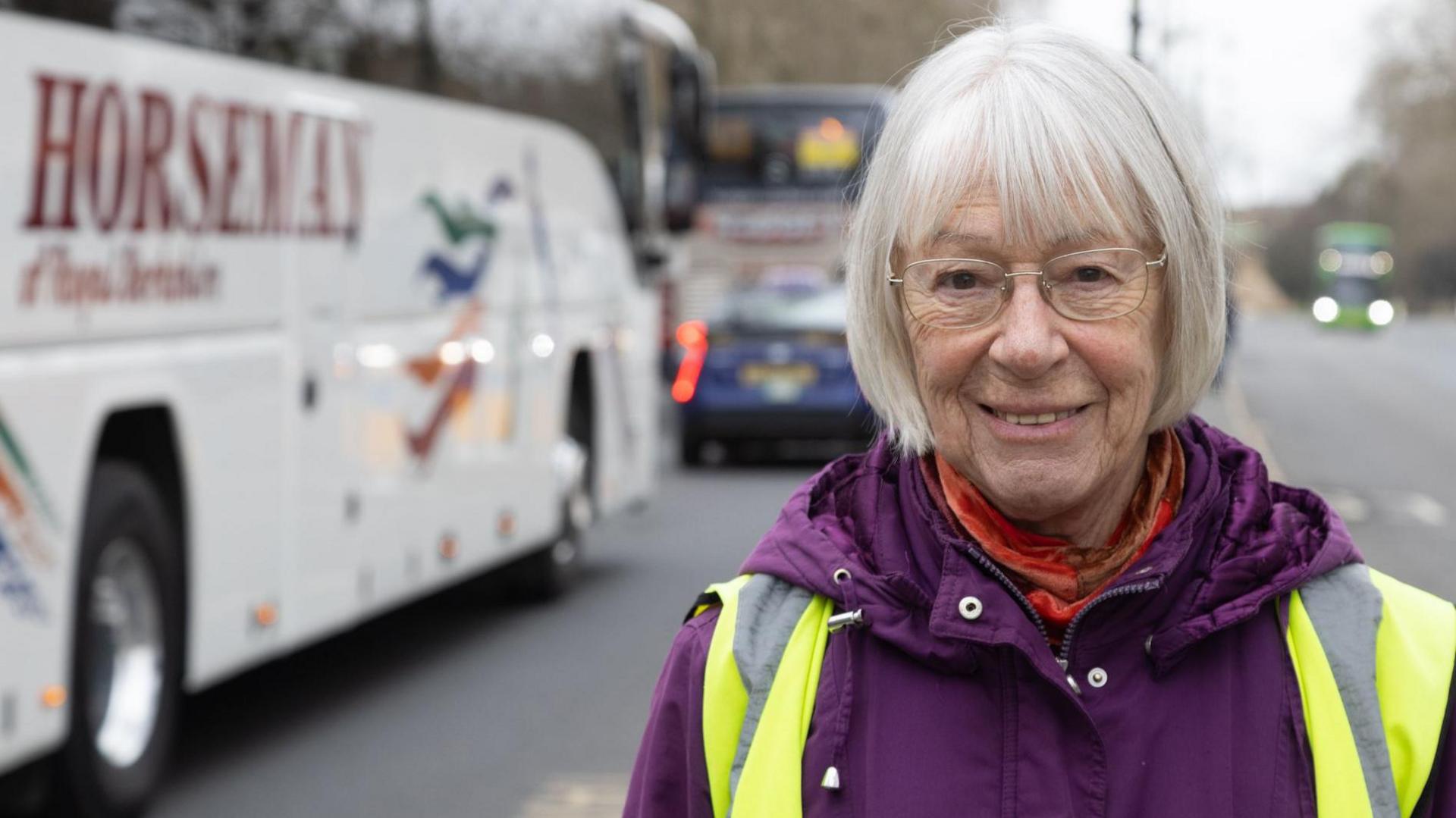 Susanna Pressel has medium length grey hair and glasses. She is wearing a purple coat and hi-vis jacket. She is stood in front of a road with a coach travelling on it.