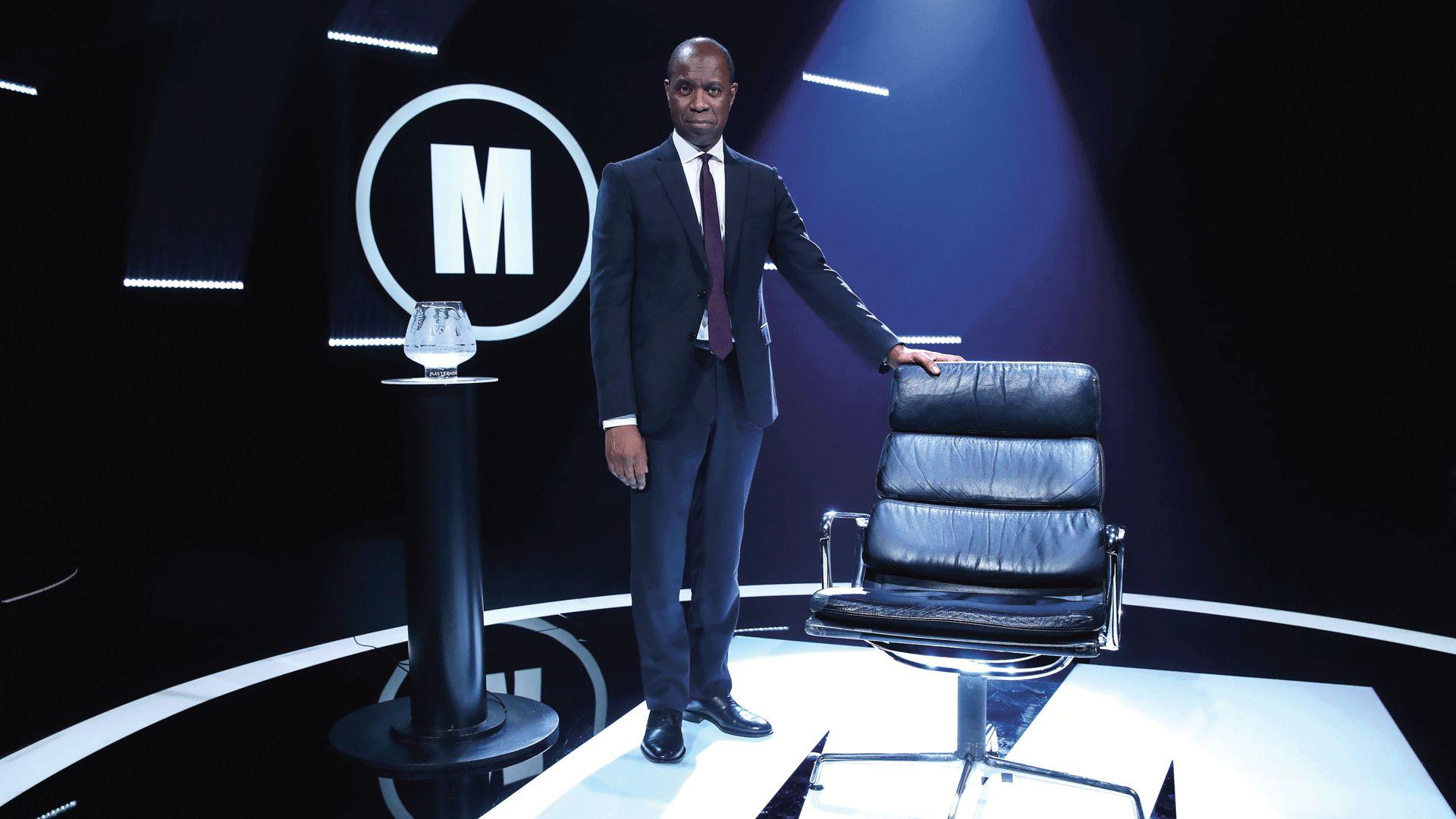 clive myrie, a man in a navy suit with a burgundy tie, stands in a black room with his right hand holding onto the top of a black leather chair. on the wall behind him is a white m in a white circle. the same logo is on the floor below clive's feet. to his left is a black podium with an etched glass bowl on it