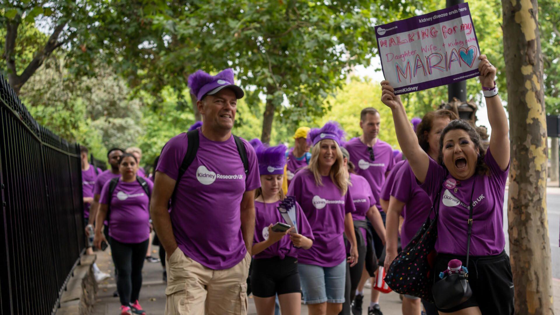 A procession of people wearing purple Kidney Research UK t-shirts walking alone a pavement. One is holding a white placard with the words "WALKING for my MARIA" written on it.