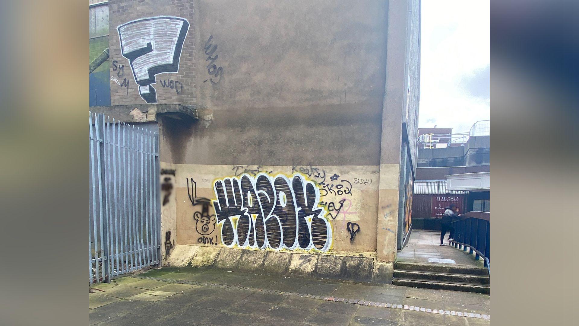 A large piece of graffiti on a wall next to an alleyway and a metal fence, close to a shopping precinct.