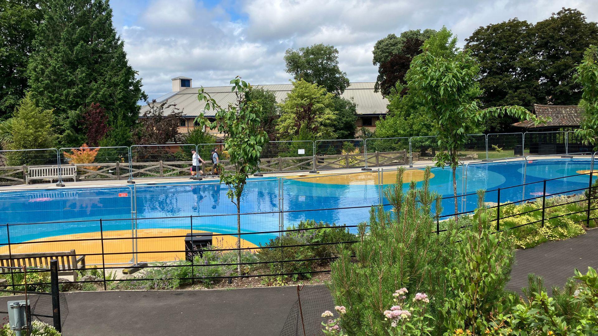 A public paddling pool with blue and yellow flooring. It is surrounded by a footpath and trees