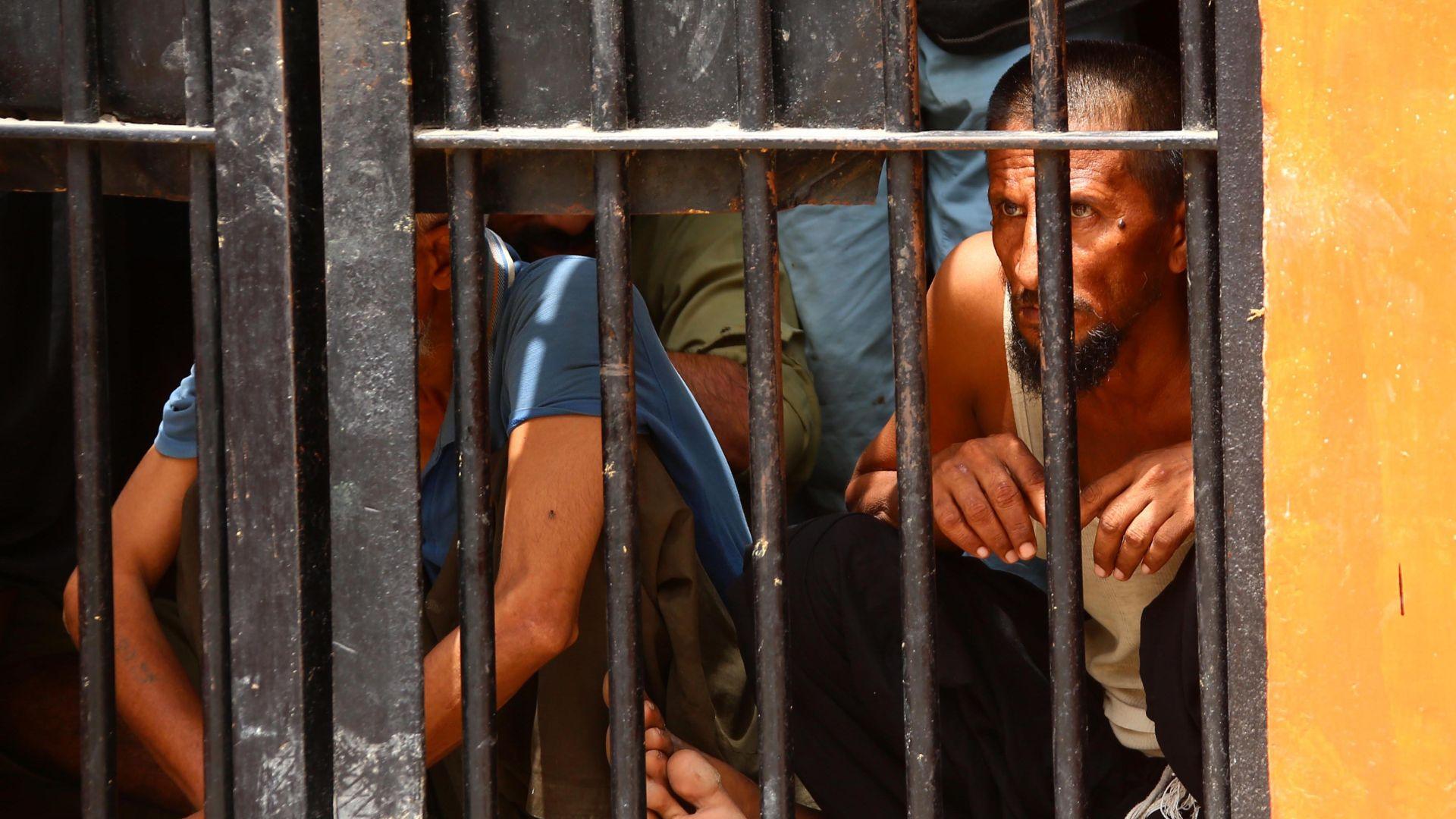 A group of men are seen crouched on the floor while behind prison cell bars.