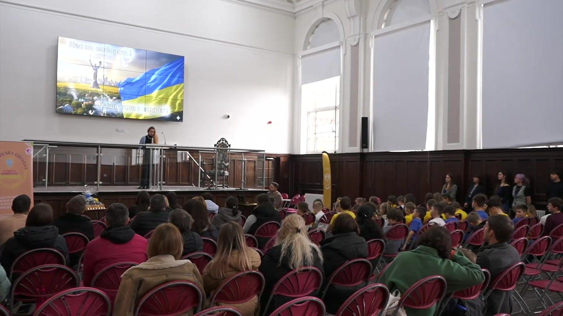 People sit in a hall on chairs and a speaker is on stage speaking to the gathered people. A monitor behind the speaker shows the flag of Ukraine. 
