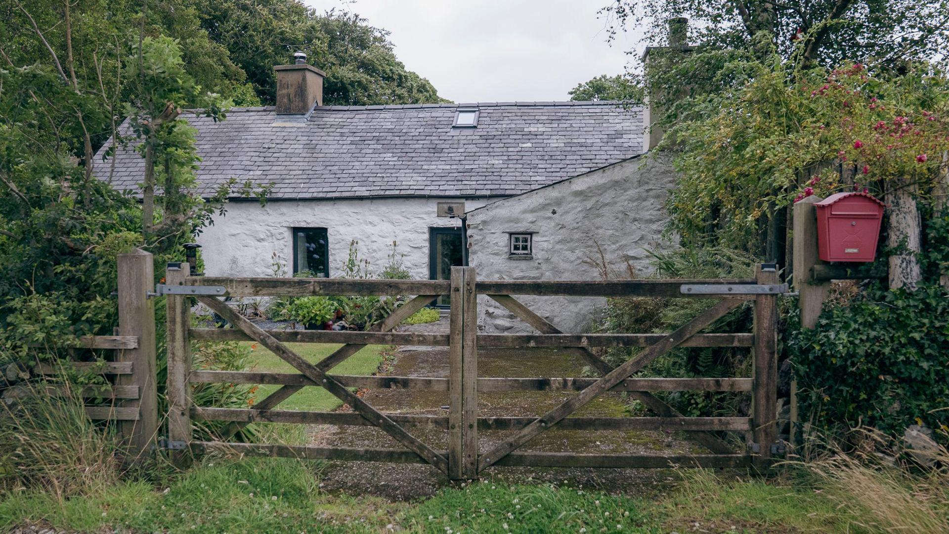 A one story white house surrounded by trees, behind a wooden gate. It has a grey roof and a red post box outside. 