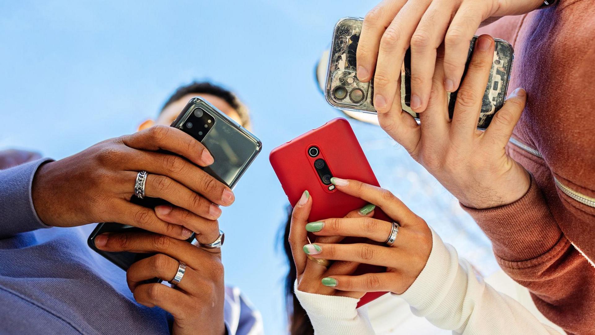 A group of three young adults stand together, all holding and looking at their own smartphone. The screens obscure their faces. One person is wearing a blue hooded jumper and has rings on their fingers. Another is wearing a white jumper with green painted nails and has a ring on their middle finger on one hand. The third is wearing a faded orange zip-up jumper. The sky is bright blue behind their heads.