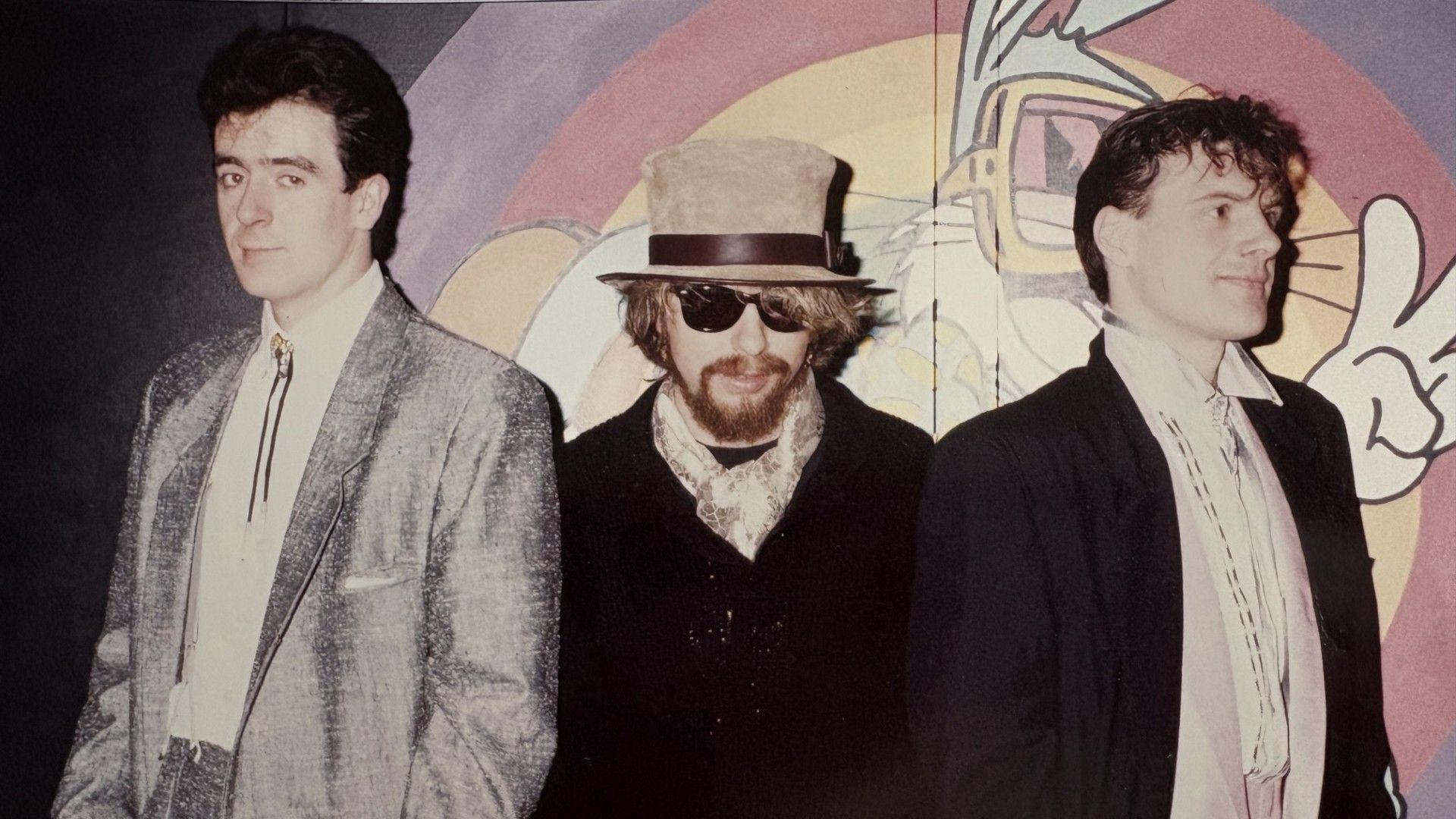 Joe Hughes (right), Dave Stewart and David Freeman, standing by a wall. Joe is looking to the camera, Dave Stewart, with glasses and a hat on, also to the camera and David to the right. A mural is behind them. 
