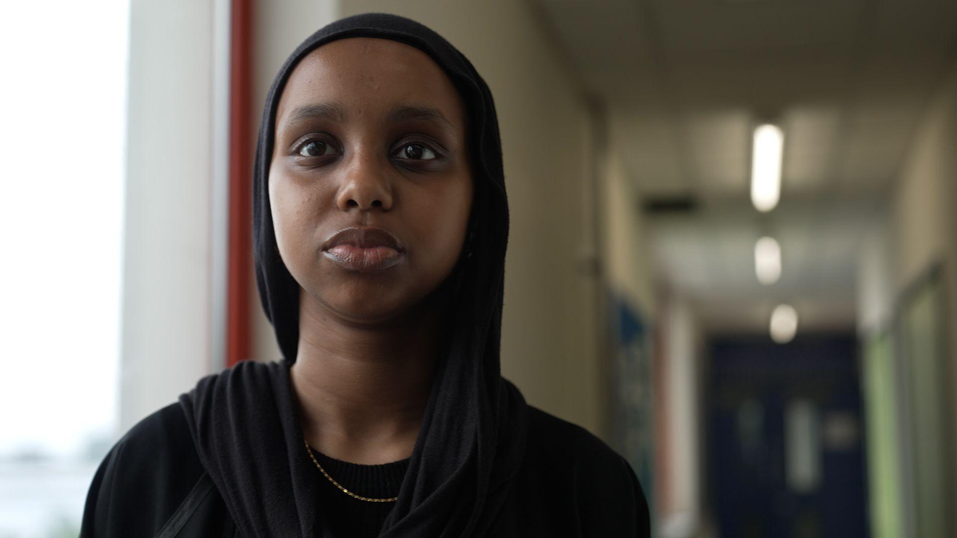 17-year-old Zaynab Ahmed is standing in a corridor near a window. She's looking at the camera. She's smartly dressed in a black head scarf, black Abaya (a floor-length dress) and a gold necklace.