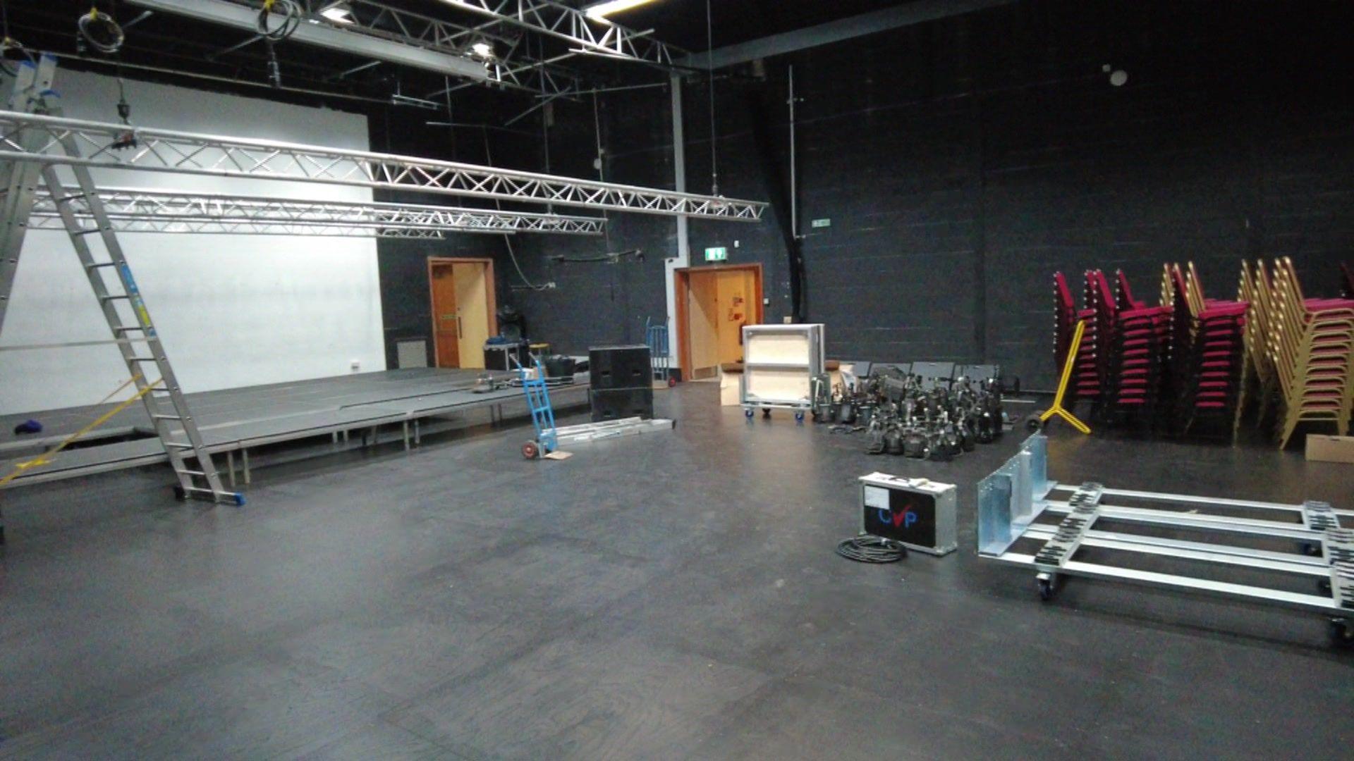 The auditorium at the mill in Banbury. On the left there is a stage and metal bars, on the right there are stacks of chairs.