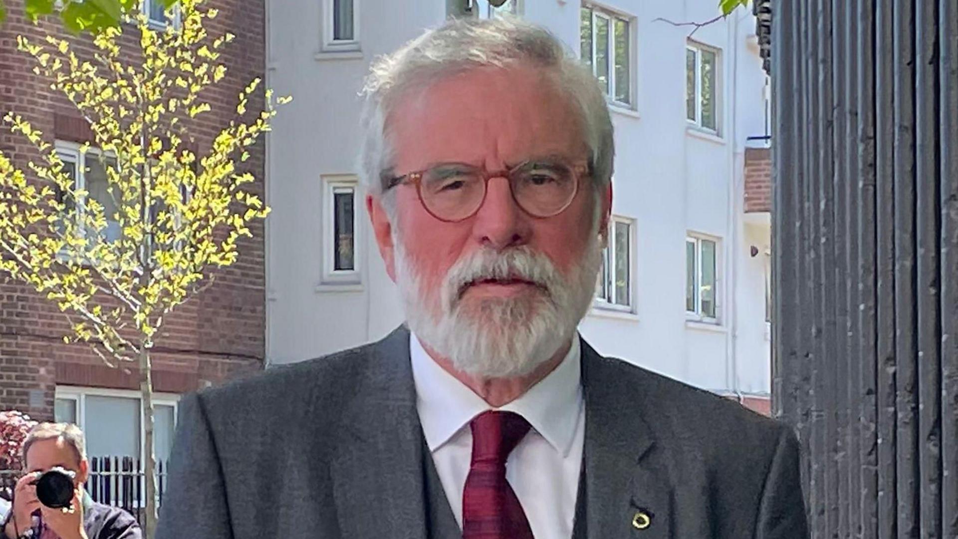 Gerry Adams is pictured from the chest up - he wears a grey jacket, grey waistcoat and red tie. He has two pins on his lapel. He has grey hair, a grey beard and brown glasses. In the background you can see a photographer taking a picture. 